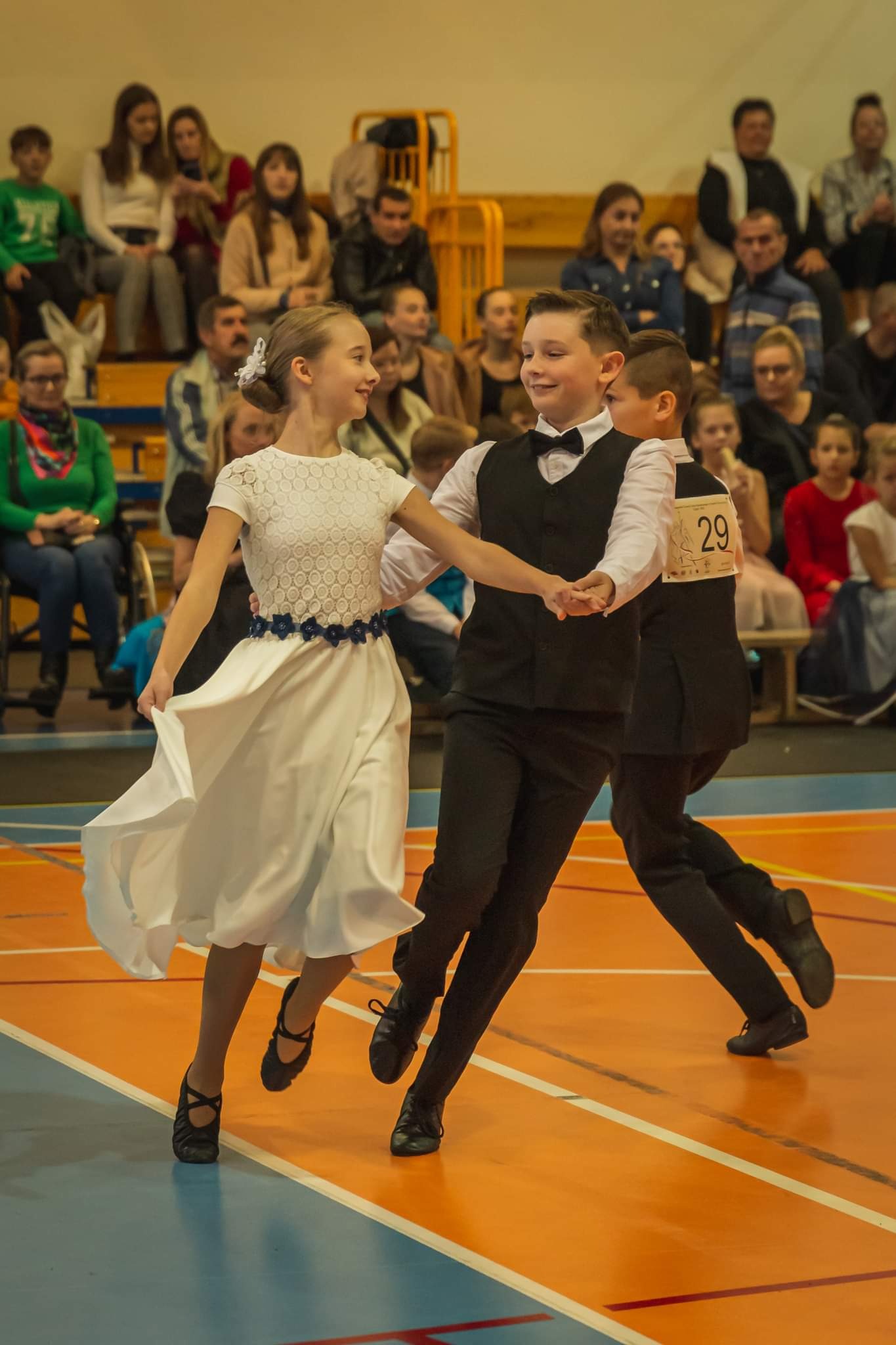Para dziecięca w strojach galowych tańczy na parkiecie podczas konkursu tańca towarzyskiego. Widzowie obserwują z trybun.