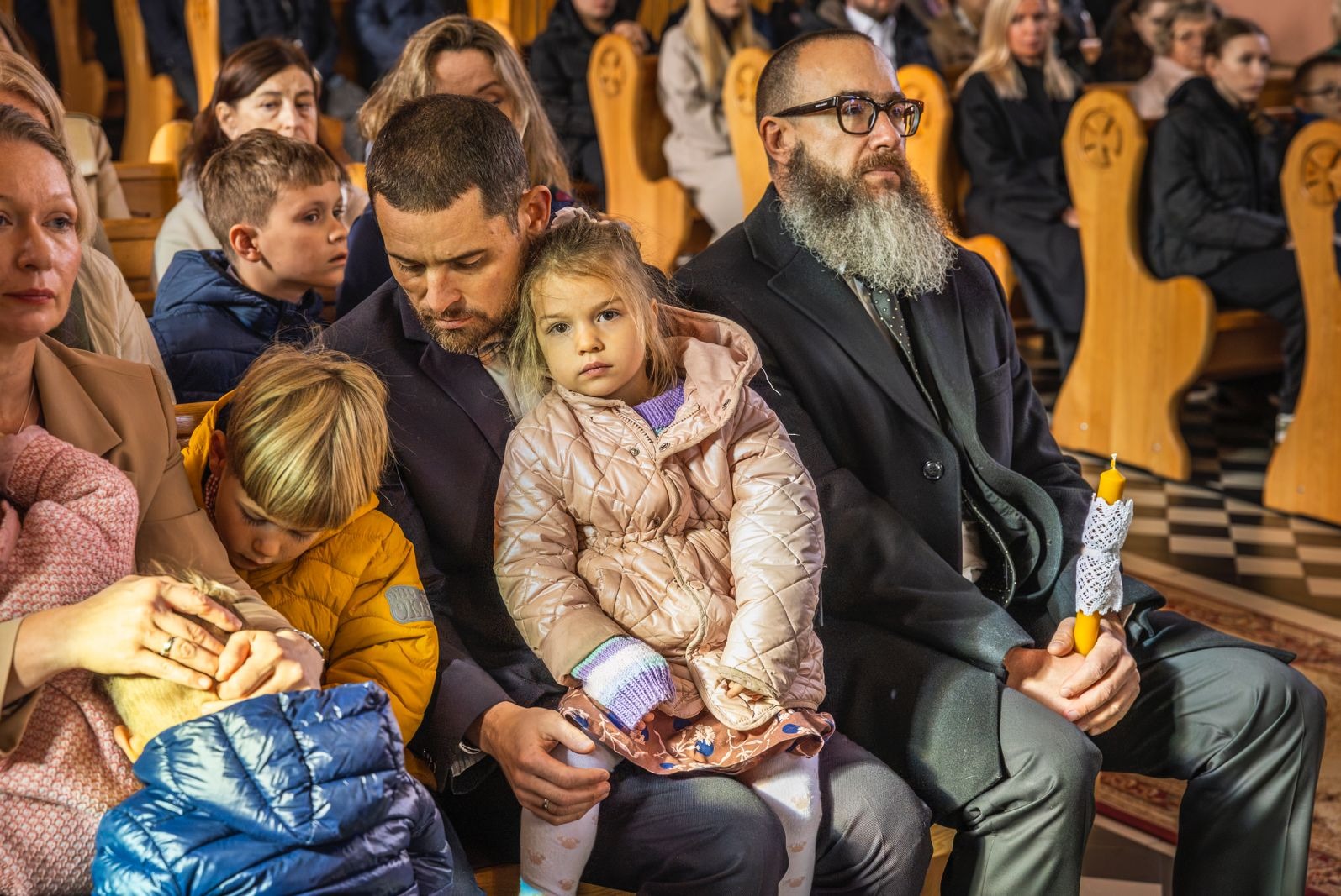Uczestnicy uroczystości w kościele: mężczyzna z brodą trzyma świecę, obok ojciec z córką na kolanach, w tle rzędy osób na drewnianych ławkach.
