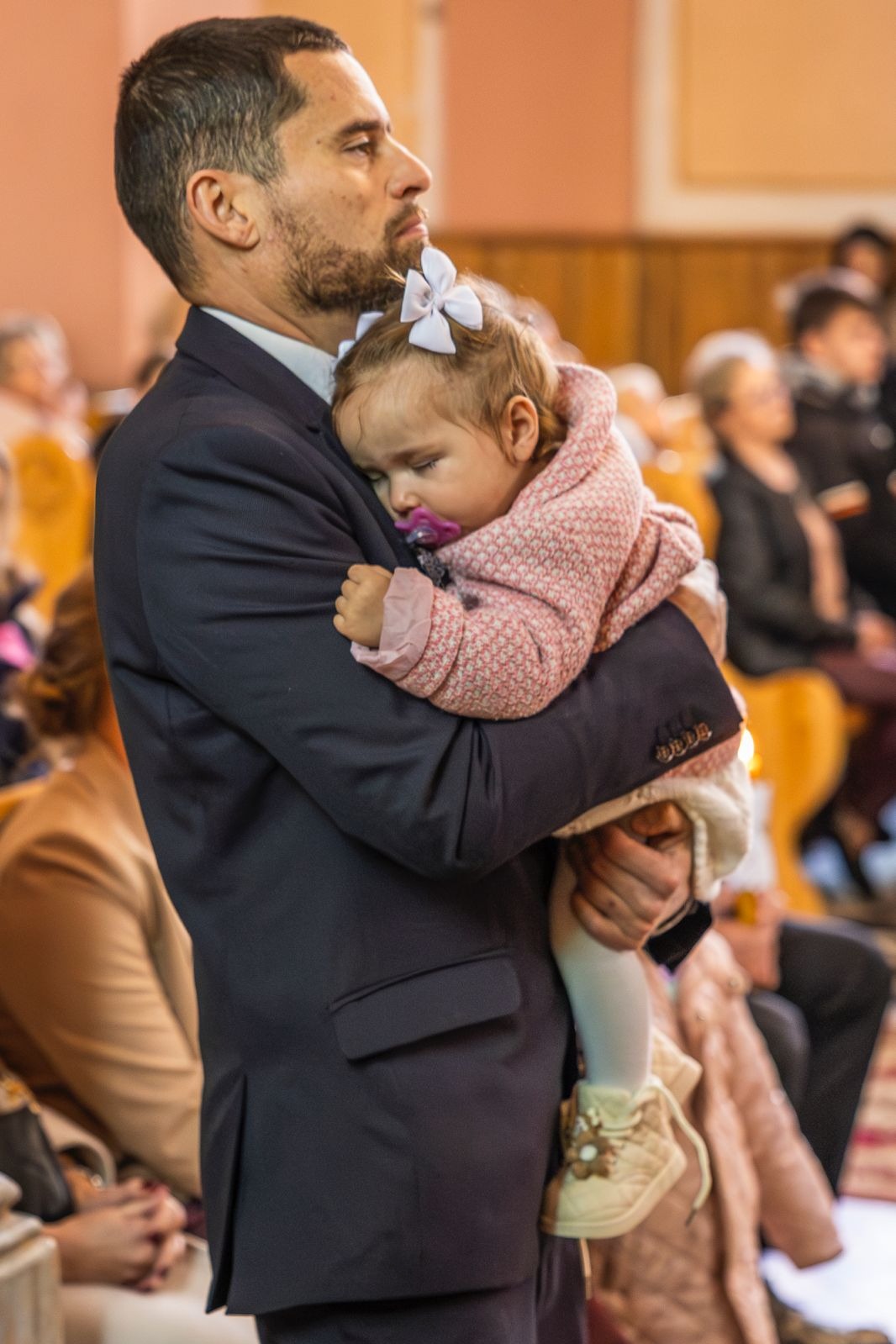 Śpiąca dziewczynka z kokardą, trzymana na rękach przez mężczyznę w garniturze, w tle widoczni ludzie, prawdopodobnie podczas uroczystości.