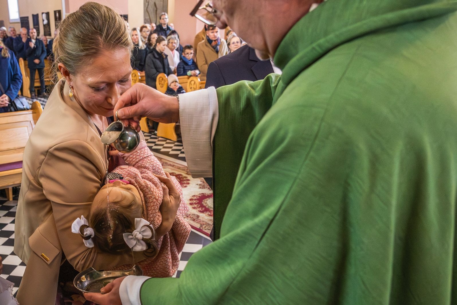 Chrzest dziecka w kościele: ksiądz polewa wodą główkę niemowlęcia trzymanego przez matkę. W tle widoczni goście, wnętrze z szachownicową podłogą i drewnianymi ławkami.