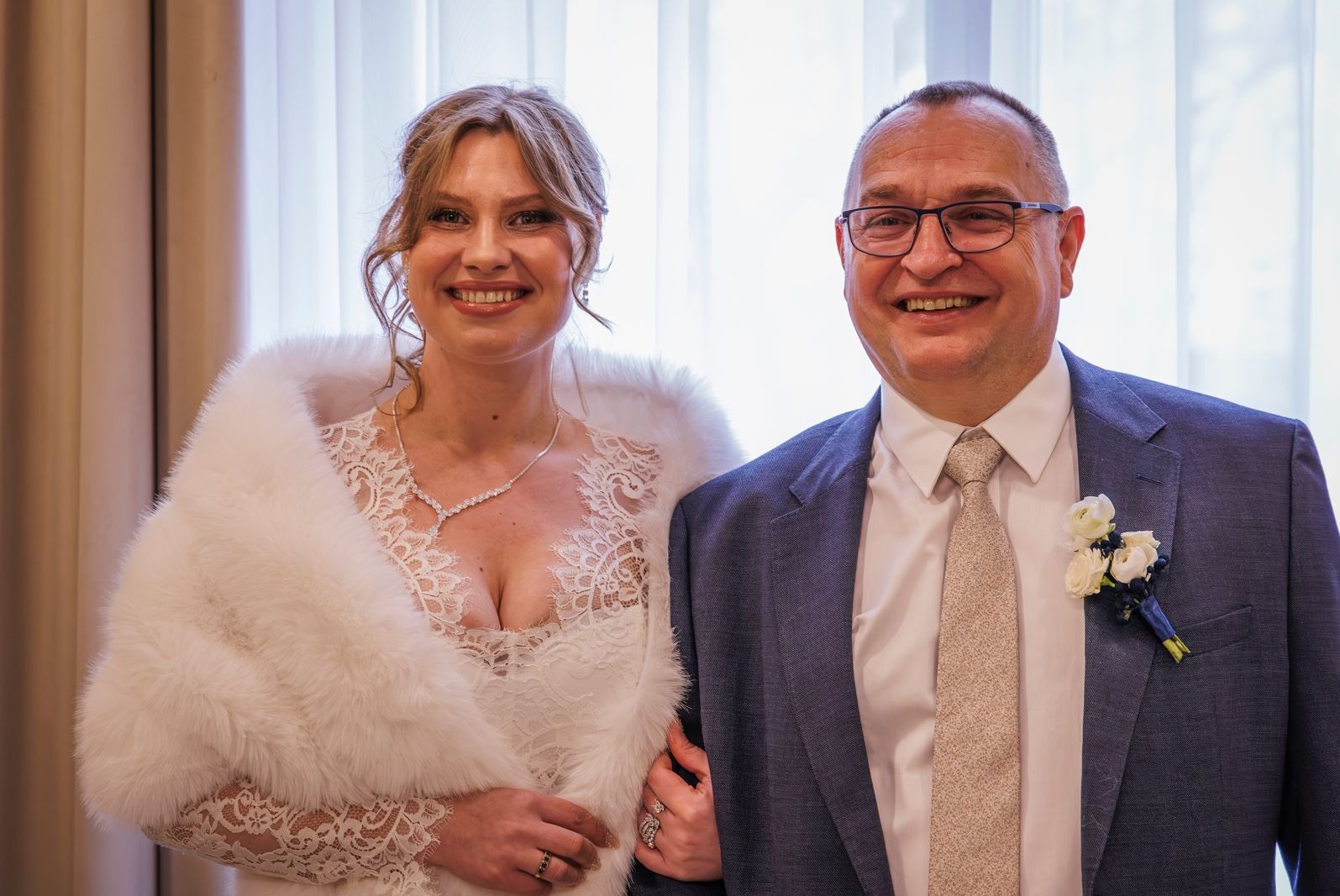 Uśmiechnięta para młoda w strojach ślubnych: panna młoda w białej sukni z futrzanym okryciem, pan młody w garniturze z butonierką. Portret w jasnym wnętrzu.