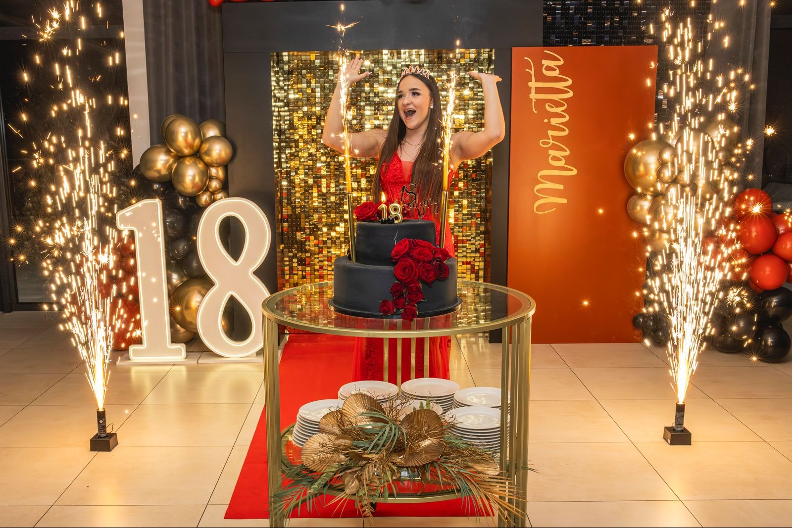 Uśmiechnięta osiemnastolatka w czerwonej sukience, z tiarą, świętuje urodziny przy czarnym torcie z różami, otoczona fontannami iskier i dekoracjami.