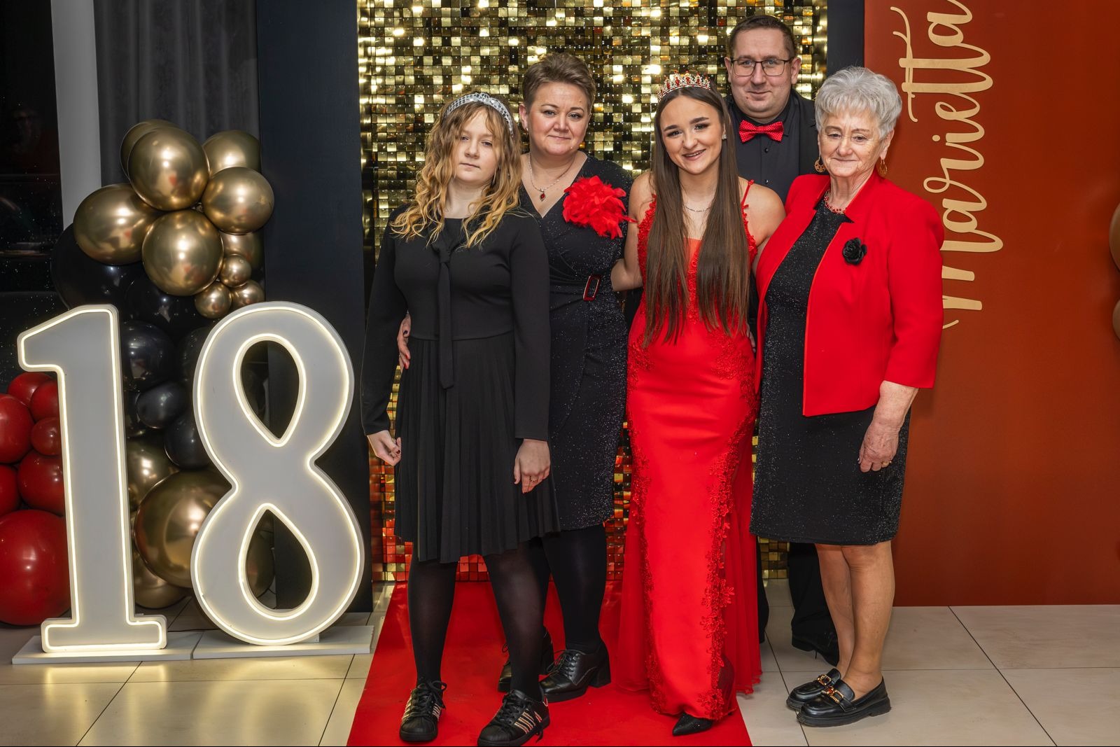 Uroczyste zdjęcie rodzinne z osiemnastki w Brzezinach. Dekoracje z balonów i błyszcząca ścianka. Dziewczyna w czerwonej sukni z koroną na głowie.