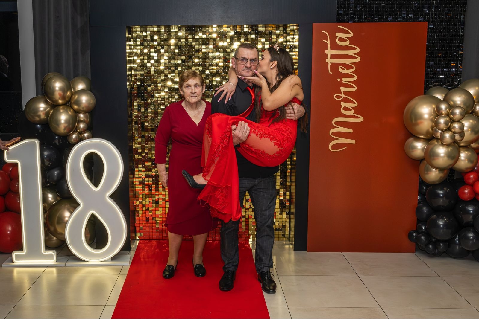 Uroczyste zdjęcie z osiemnastki: mężczyzna trzyma na rękach całowaną przez kobietę w koronie, obok stoi starsza pani, tło z cekinami i balonami.