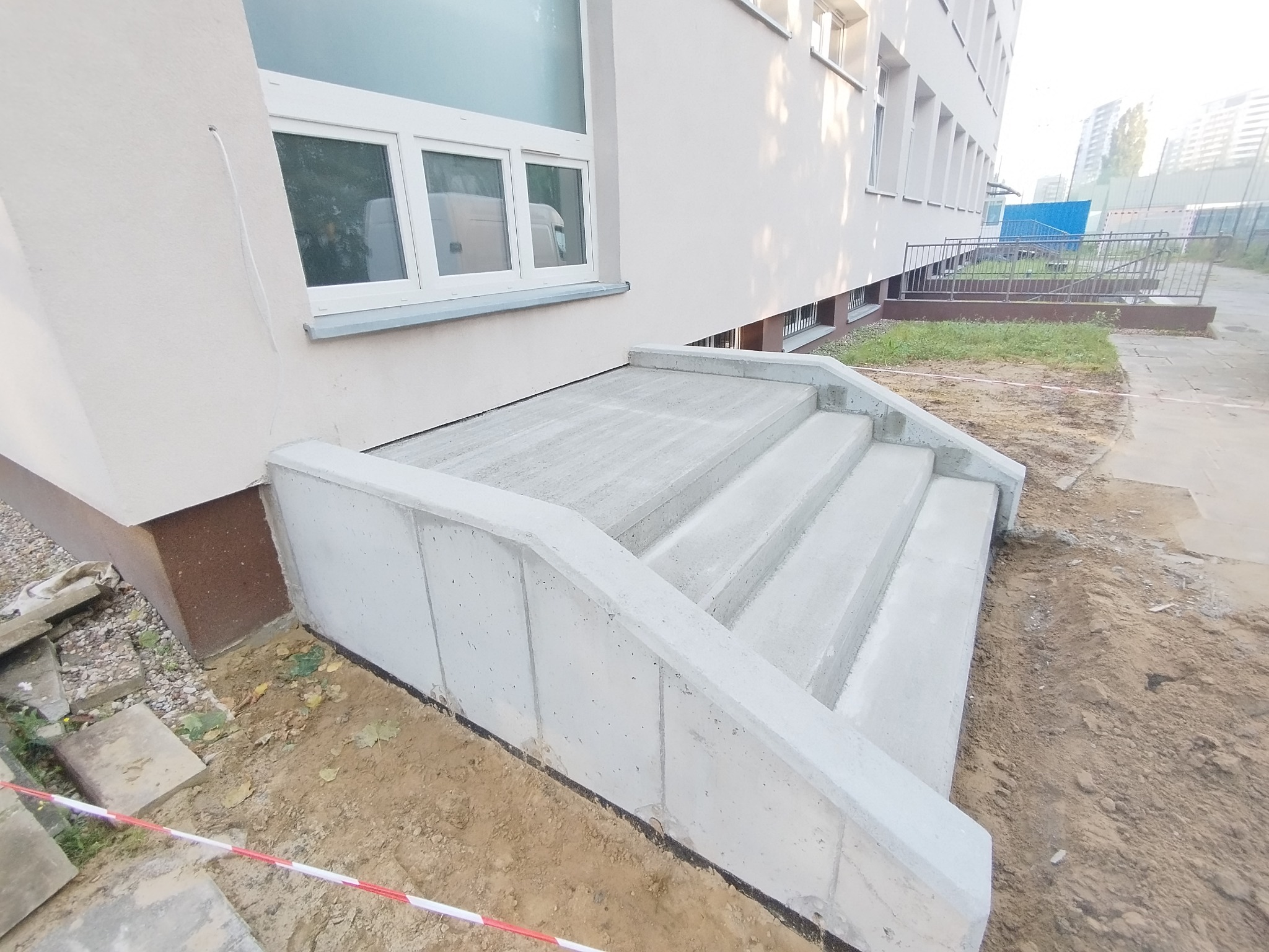 Nowe, betonowe schody wejściowe do budynku, z widoczną strukturą betonu i świeżo uporządkowanym terenem wokół. Ujęcie z niskiej perspektywy podkreśla solidność konstrukcji.