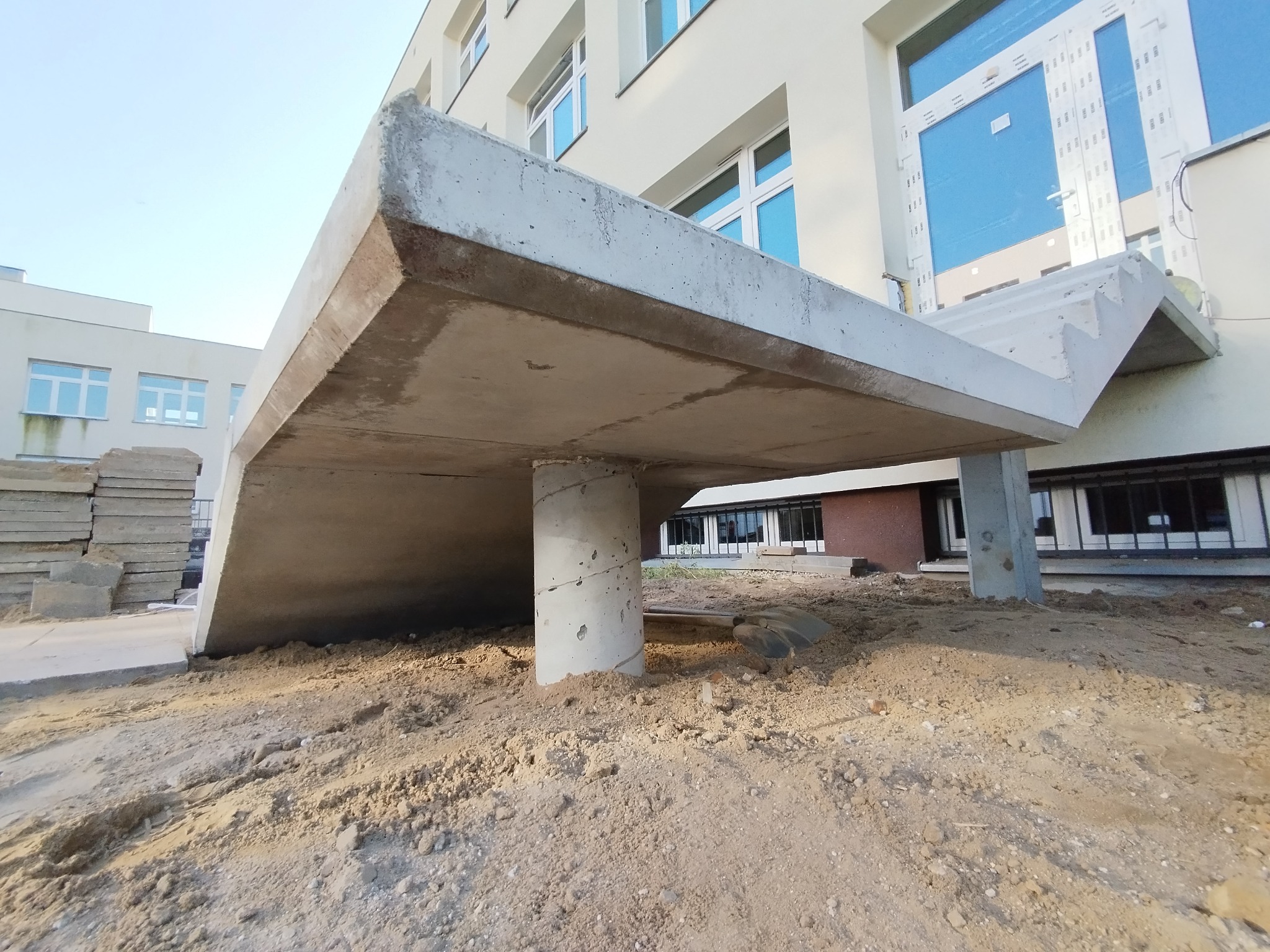 Surowy betonowy strop podtrzymywany przez kolumny na placu budowy, z widoczną łopatą i stertą płyt chodnikowych w tle budynku z oknami.