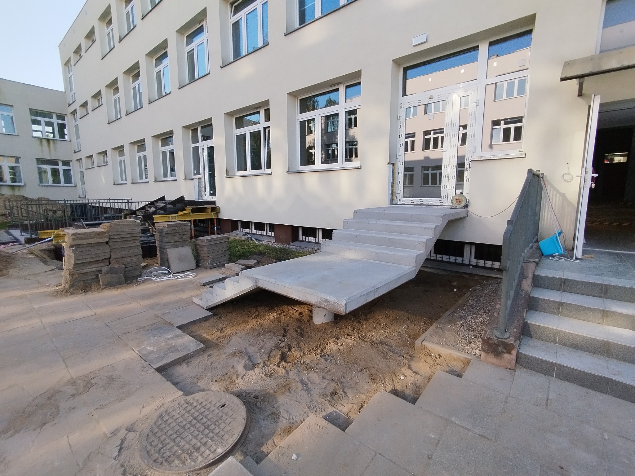 Betonowe schody wejściowe do budynku w trakcie budowy, z widocznymi elementami konstrukcyjnymi i przygotowanym terenem. Widok na elewację budynku z oknami.