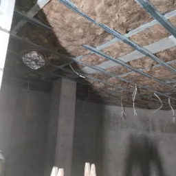 Montaż izolacji z wełny mineralnej na poddaszu, widoczne metalowe profile sufitowe i wystające przewody elektryczne, zabezpieczony folią otwór wentylacyjny.