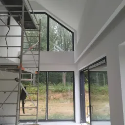 Wnętrze domu w trakcie malowania, widoczne rusztowanie, duże okna z czarnymi ramami i pomalowane na biało ściany.