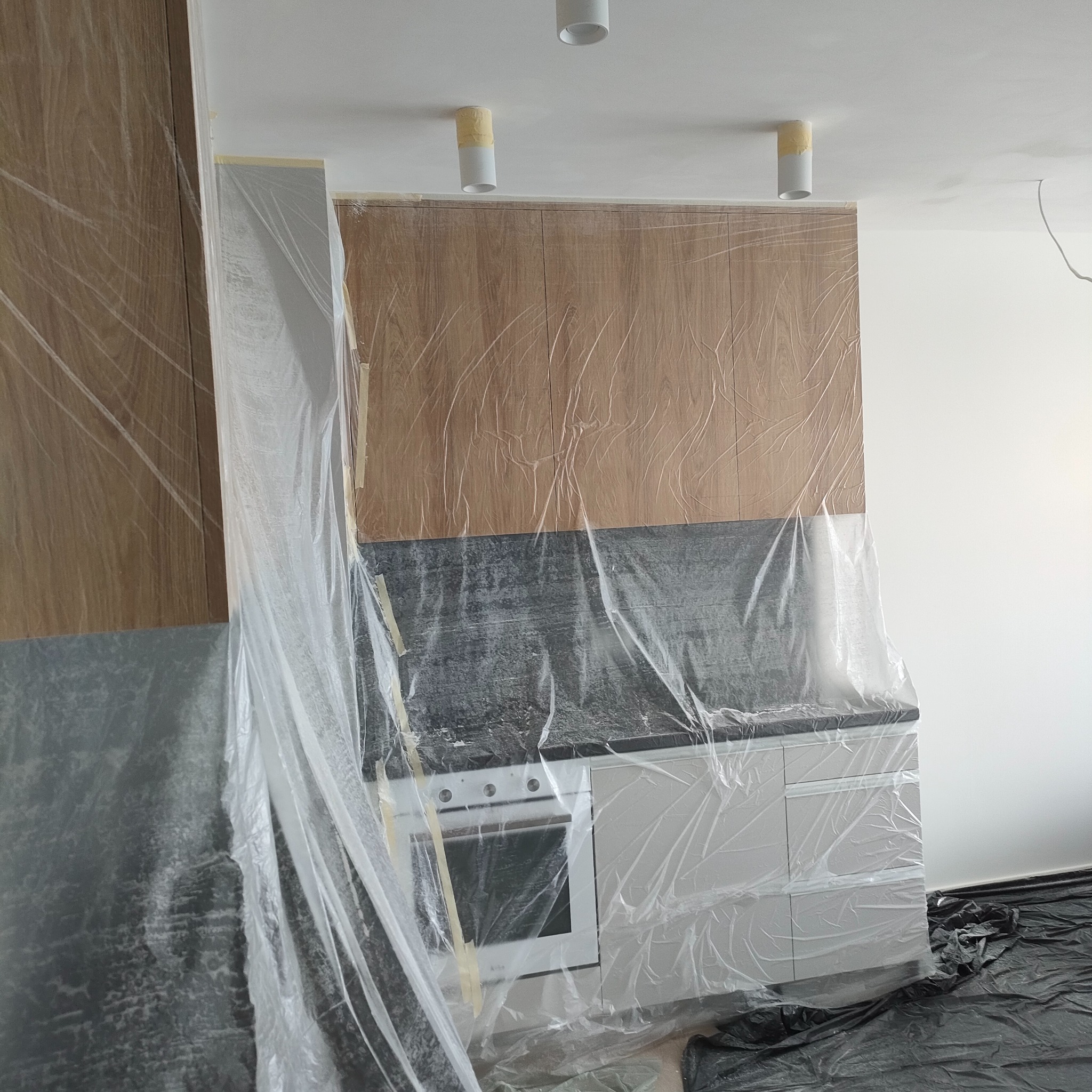 Kuchnia zabezpieczona folią malarską podczas prac remontowych; widoczne zaklejone taśmą malarską lampy sufitowe, meble kuchenne i podłoga przykryte folią ochronną.