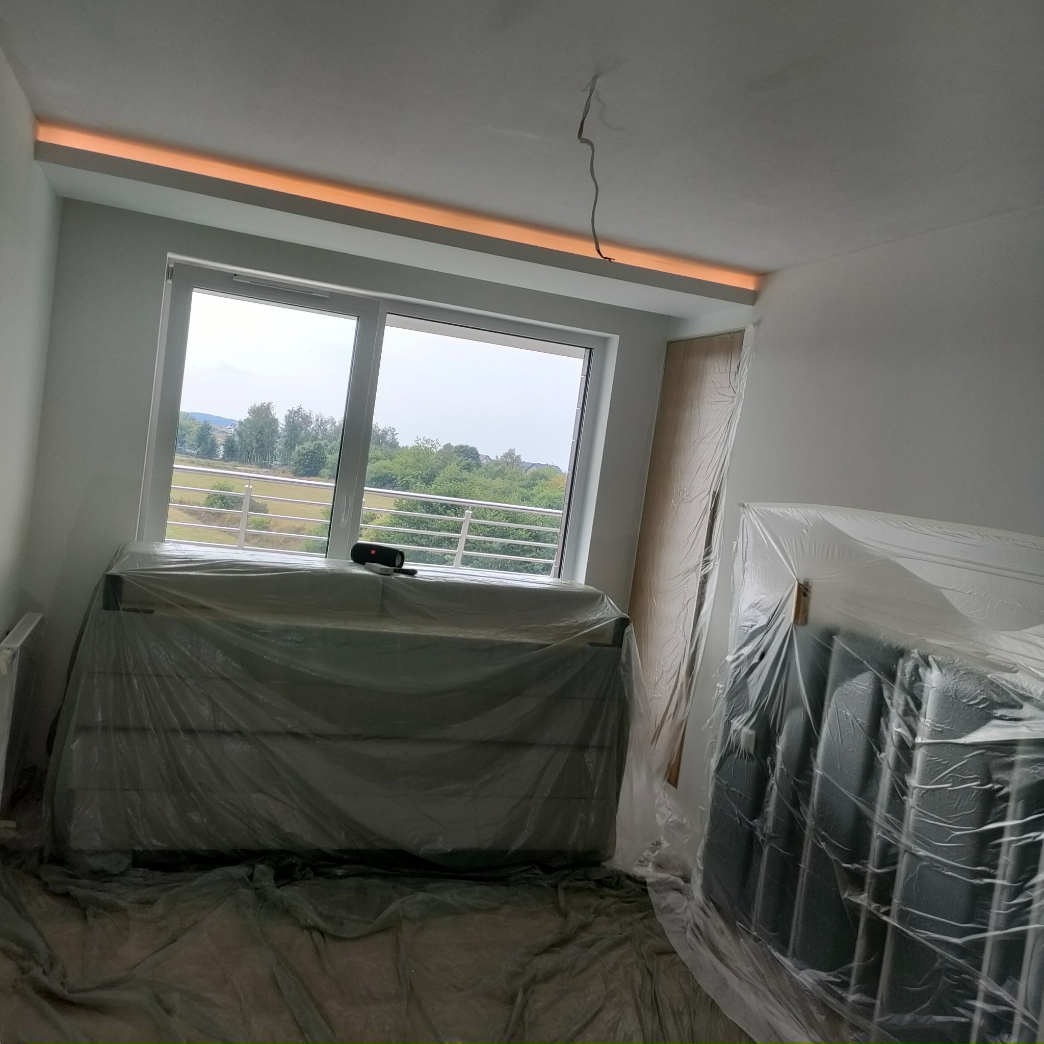 Pokój w trakcie prac wykończeniowych: okno z widokiem na zieleń, grzejniki i meble zabezpieczone folią, listwa oświetleniowa pod sufitem, ślady po szpachlowaniu na ścianach.