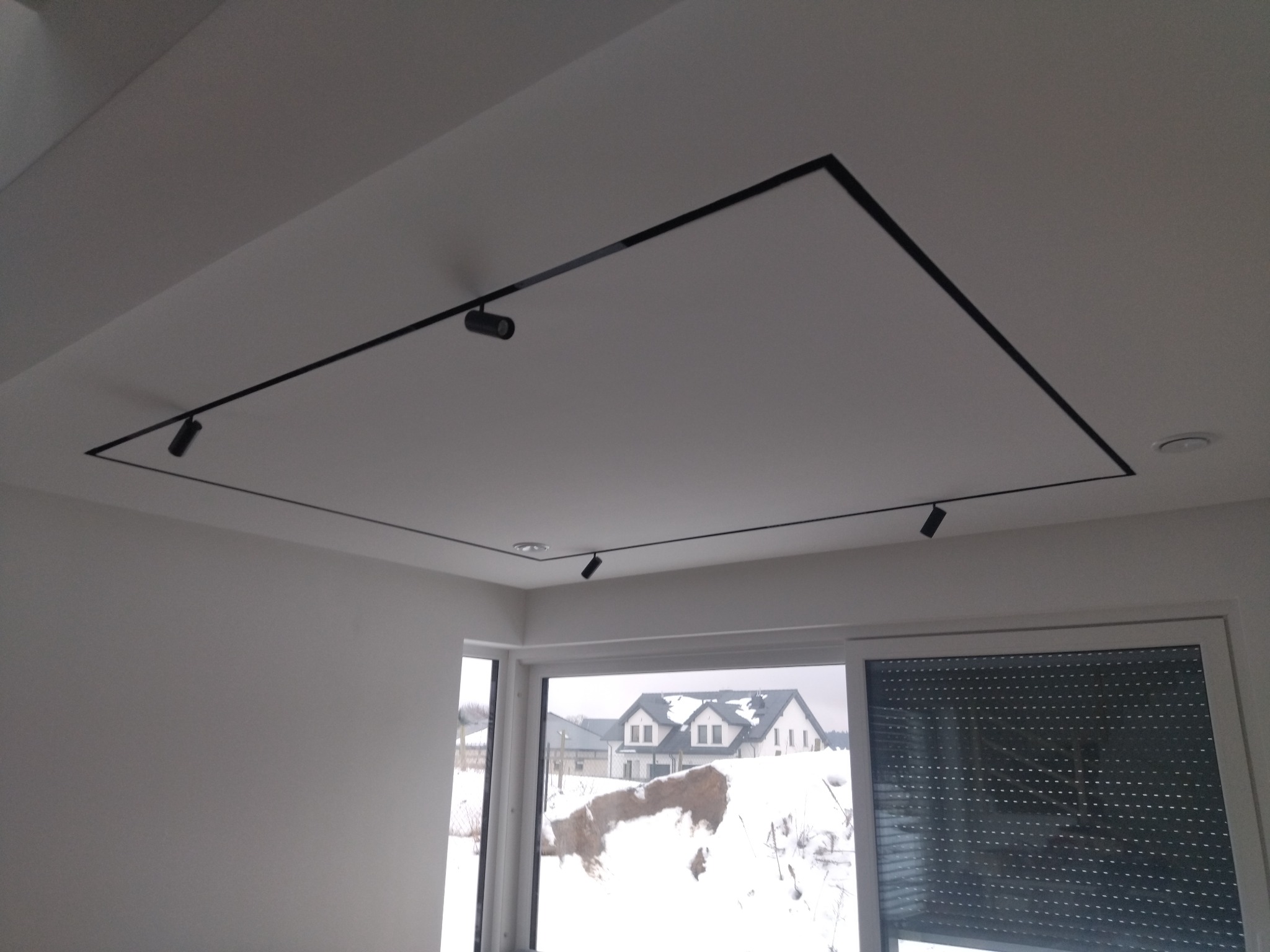 Nowoczesne oświetlenie szynowe z reflektorami w kształcie walców, zamontowane na suficie podwieszanym w kształcie prostokąta z czarną obwódką, widok na okno z roletą i ośnieżony krajobraz za oknem.