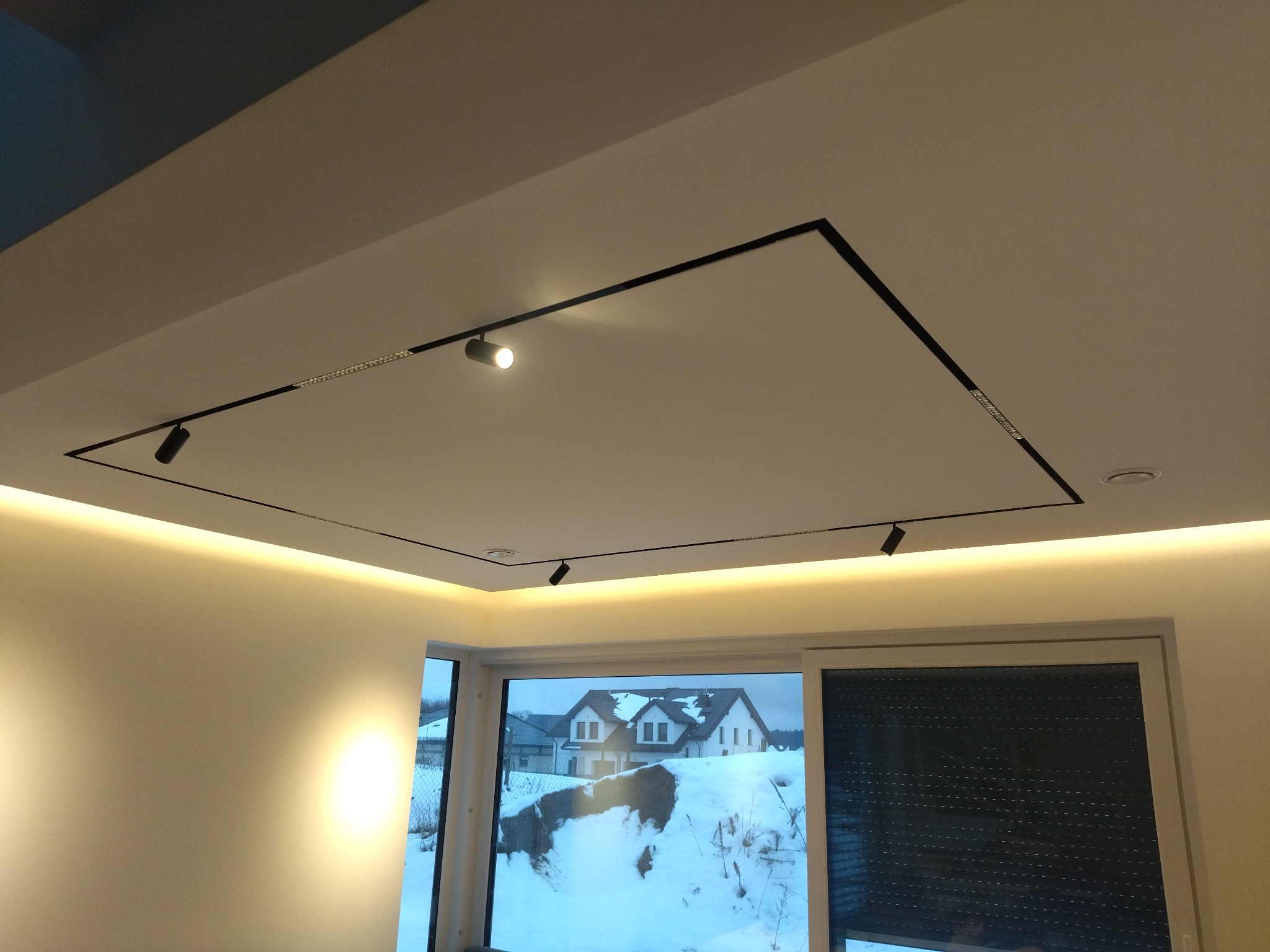 Nowoczesne oświetlenie sufitowe z czarnymi szynami i punktowymi reflektorami w białym wnętrzu, widok na zaśnieżony krajobraz za oknem.