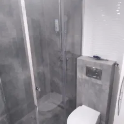 Wykończona łazienka z szarą glazurą, kabina prysznicowa z przezroczystego szkła, biała toaleta podwieszana, na obudowie stelaża narzędzia.