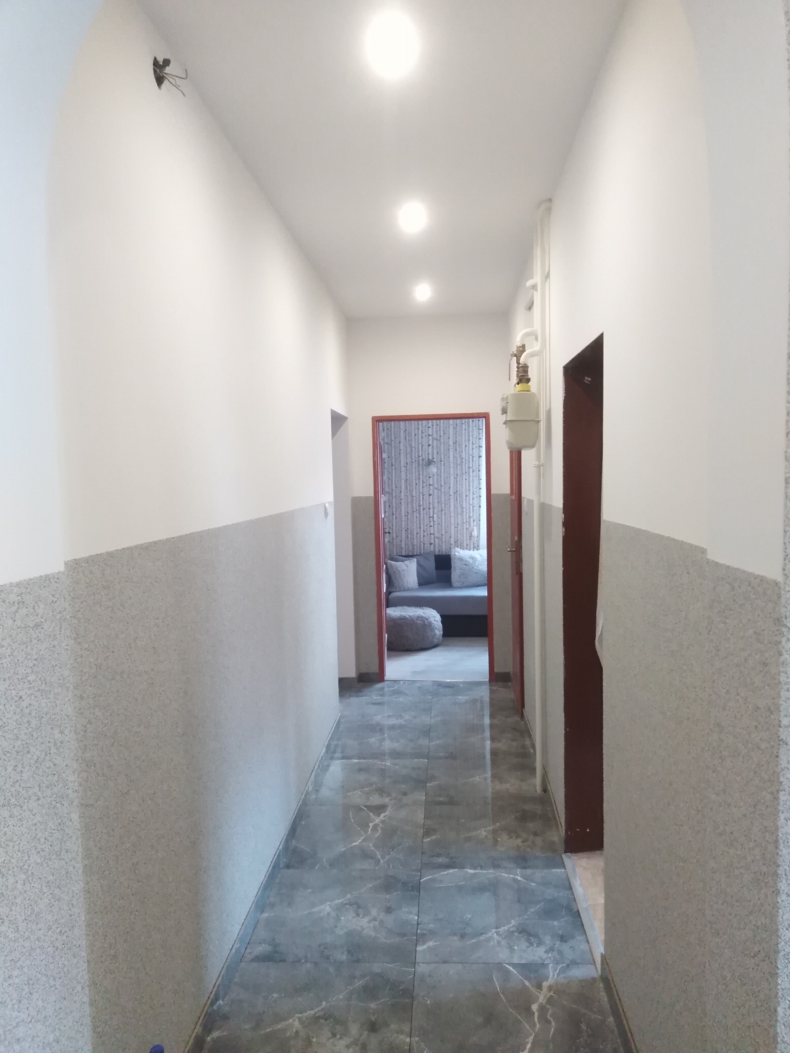 Korytarz po remoncie z szarymi płytkami imitującymi marmur na podłodze i ścianach do połowy wysokości, białe ściany powyżej, widoczny gazomierz i fragment salonu w tle.