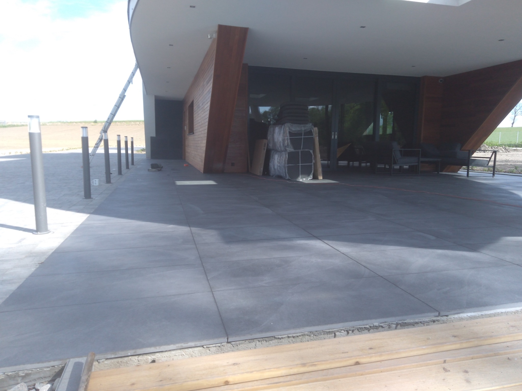 Szare płytki chodnikowe ułożone na zewnątrz budynku z drewnianą elewacją, widoczne materiały budowlane i drabina.