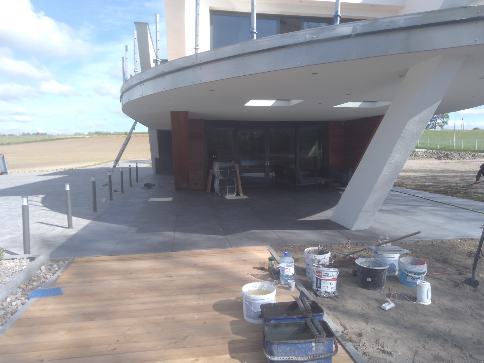 Nowoczesny dom w trakcie wykańczania: świeżo położona drewniana podłoga tarasowa, widoczne wiadra z zaprawą, narzędzia i czajnik elektryczny, na zewnątrz polne tereny.