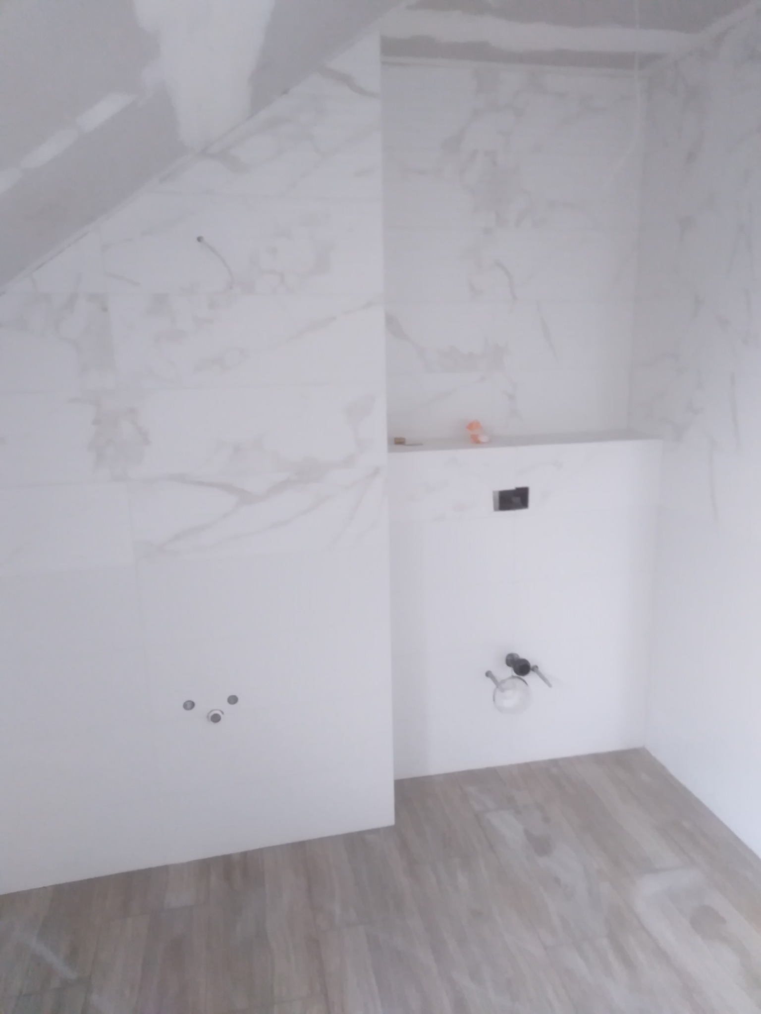 Wnętrze łazienki w trakcie prac wykończeniowych; widoczne płytki imitujące marmur na ścianach, drewnopodobne panele na podłodze, przygotowane punkty instalacji wodnej, podwieszana półka i zabudowa...