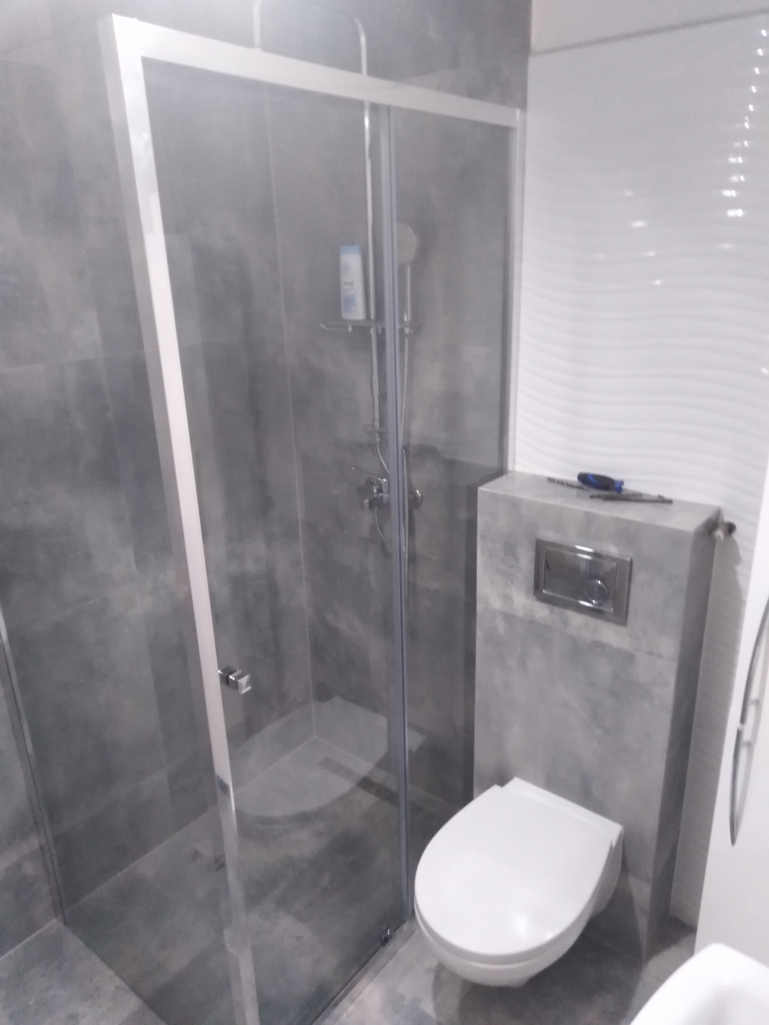 Wykończona łazienka z szarą glazurą, kabina prysznicowa z przezroczystego szkła, biała toaleta podwieszana, na obudowie stelaża narzędzia.