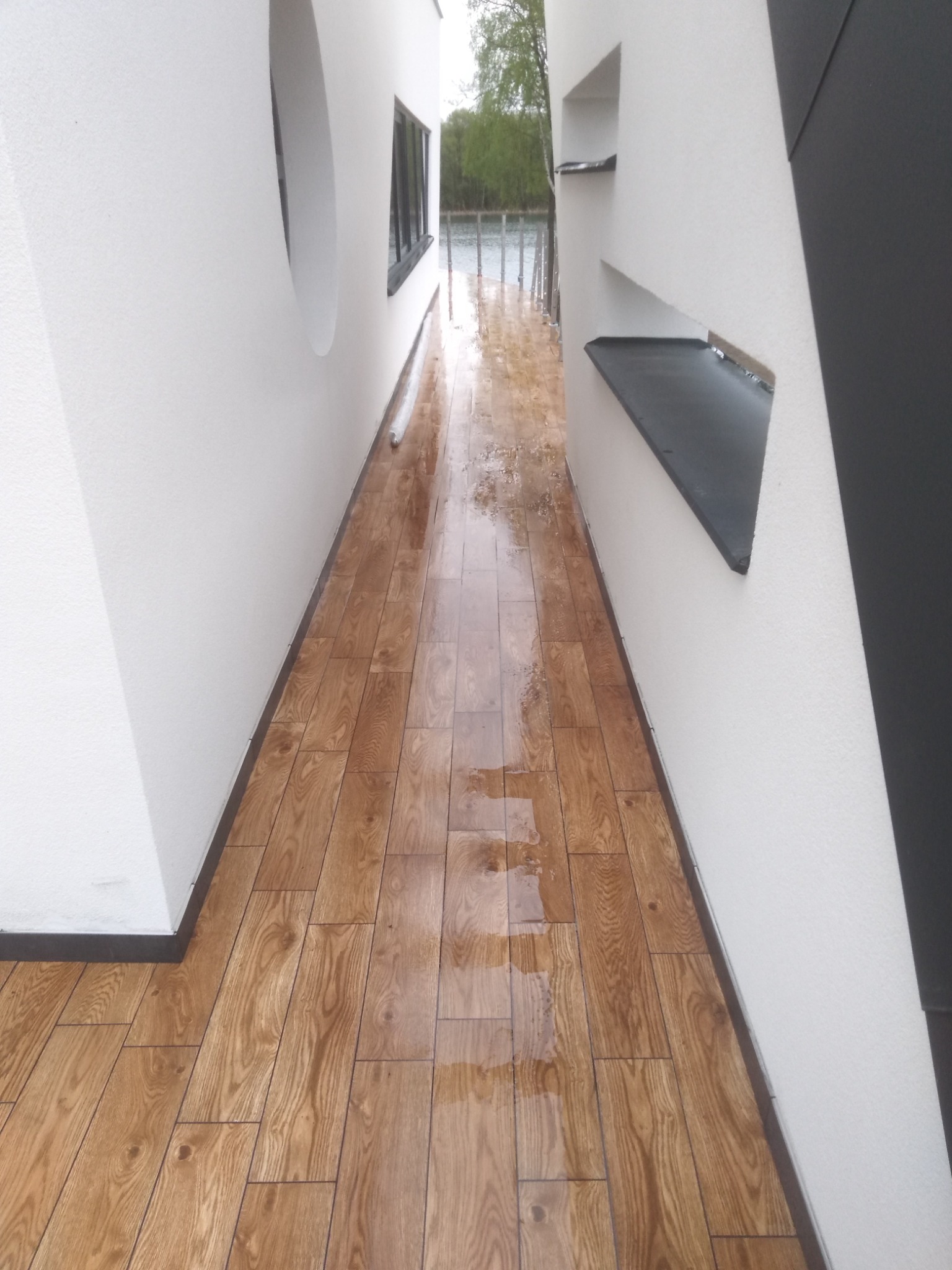 Wąski korytarz z drewnopodobną podłogą z płytek, białe ściany z wnękami i okrągłym oknem, widok na zewnątrz na jezioro w pochmurny dzień.