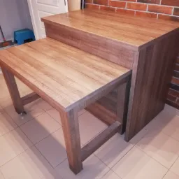 Blat drewniany zamontowany na szafce, tworzący dwupoziomowy stolik roboczy w kuchni, z widoczną teksturą drewna i ceglaną ścianą w tle.