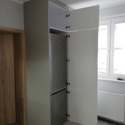 FRIZAR Meble Ryszard Kuchciński - Szara szafa wnękowa z otwartymi drzwiami, wewnątrz widoczna lodówka, na podłodze panele, obok drewniane drzwi i biały grzejnik pod oknem.