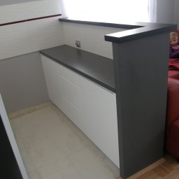 FRIZAR Meble Ryszard Kuchciński - Nowoczesna kuchnia z blatem w kolorze grafitu i białymi szafkami w Warszawie, widok na fragment salonu z czerwoną sofą i dziewczynką siedzącą na niej.