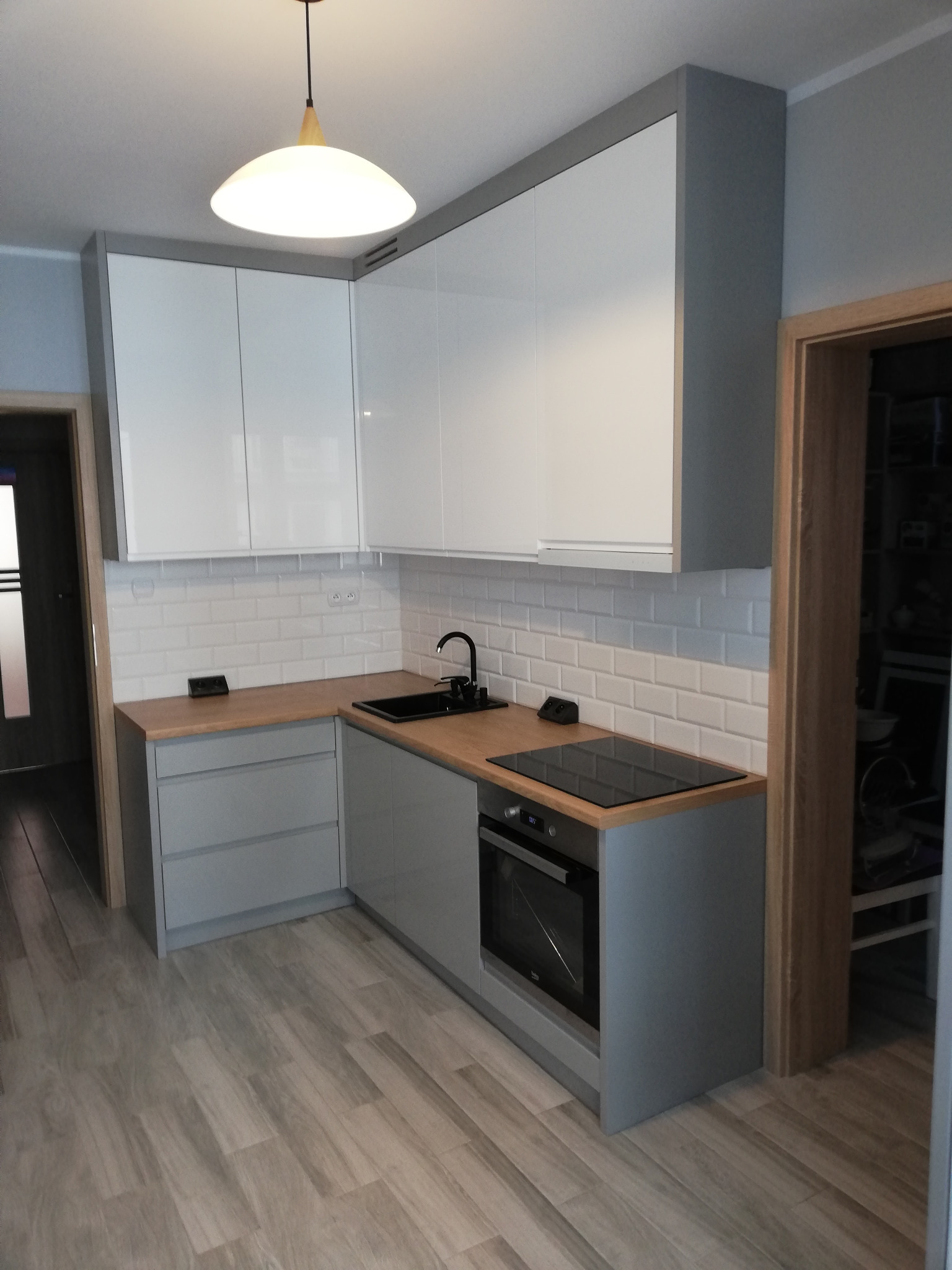 Nowoczesna, narożna kuchnia w odcieniach szarości i bieli z drewnianym blatem, czarnym zlewem i płytą indukcyjną; widoczne białe, prostokątne płytki na ścianie i lampa sufitowa w stylu skandynawskim.
