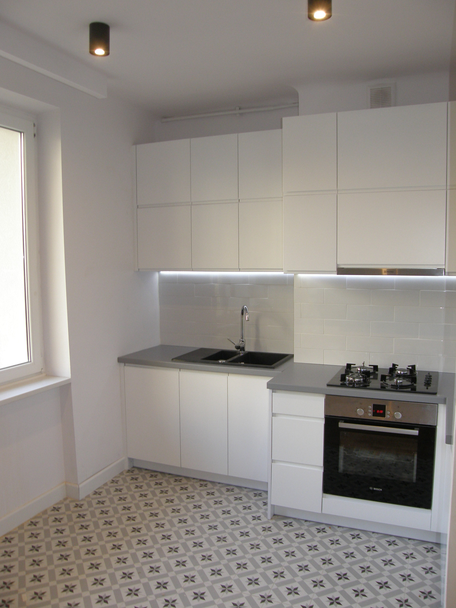 Nowoczesna, jasna kuchnia z białymi frontami szafek, szarym blatem i wzorzystą podłogą. Widoczny zlew z czarną komorą, płyta gazowa i piekarnik Bosch. Oświetlenie podszafkowe LED.