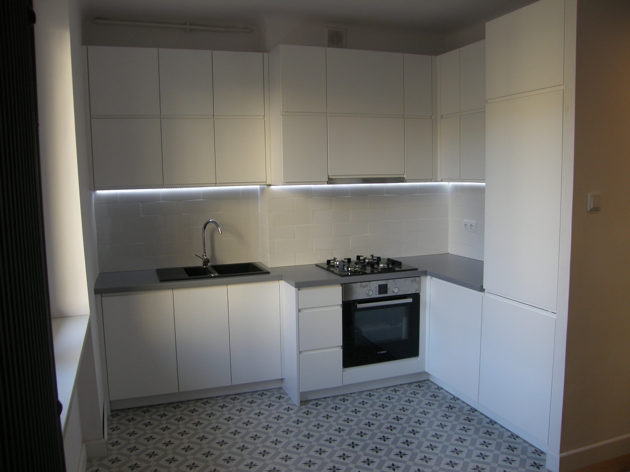 Jasna kuchnia z białymi frontami, szarym blatem i wzorzystą podłogą. Oświetlenie LED pod szafkami wiszącymi. Widoczna płyta gazowa i piekarnik.