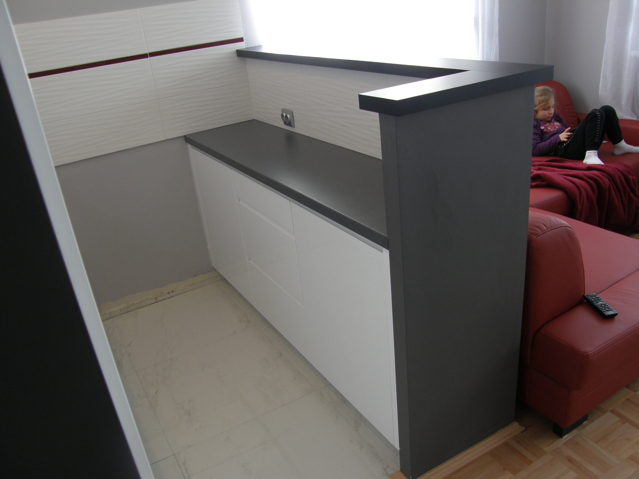 Nowoczesna kuchnia z blatem w kolorze grafitu i białymi szafkami w Warszawie, widok na fragment salonu z czerwoną sofą i dziewczynką siedzącą na niej.