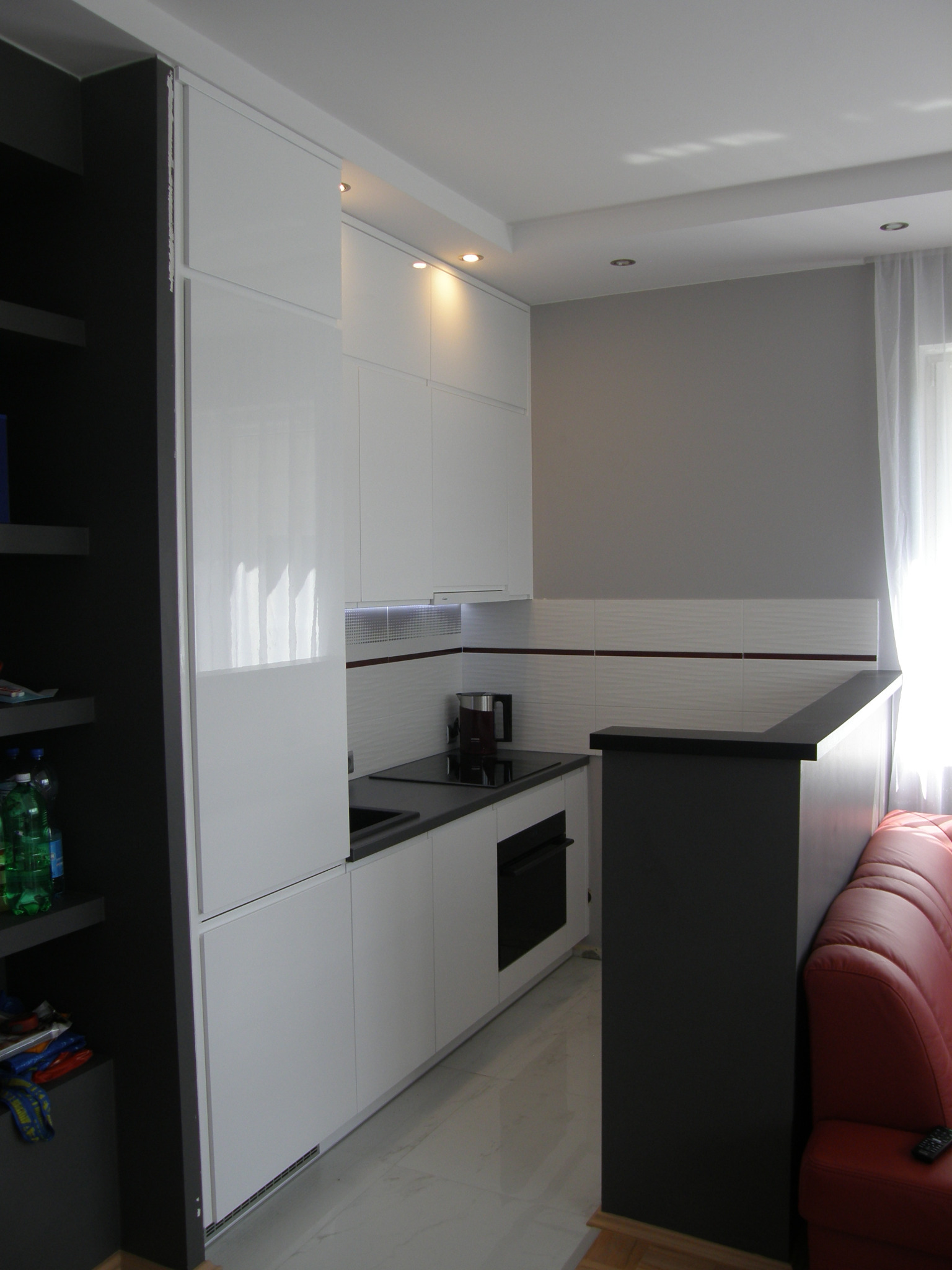 Nowoczesna, biała zabudowa kuchenna z czarnym blatem, oświetleniem LED podszafkowym i czarnym barkiem oddzielającym kuchnię od salonu, widoczny fragment czerwonej sofy.