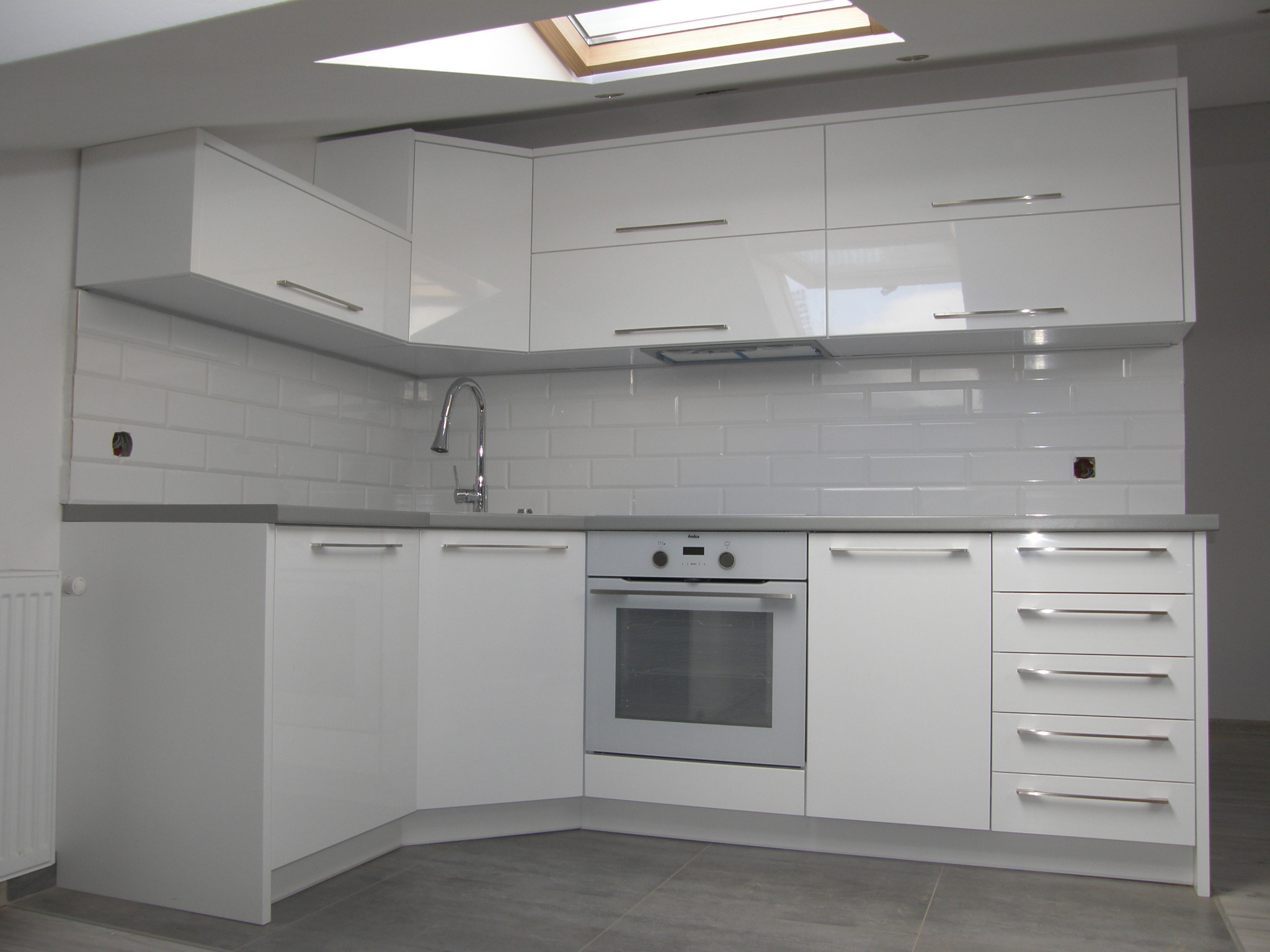 Nowoczesna, biała kuchnia z połyskiem, szary blat, białe płytki na ścianie i okno dachowe.