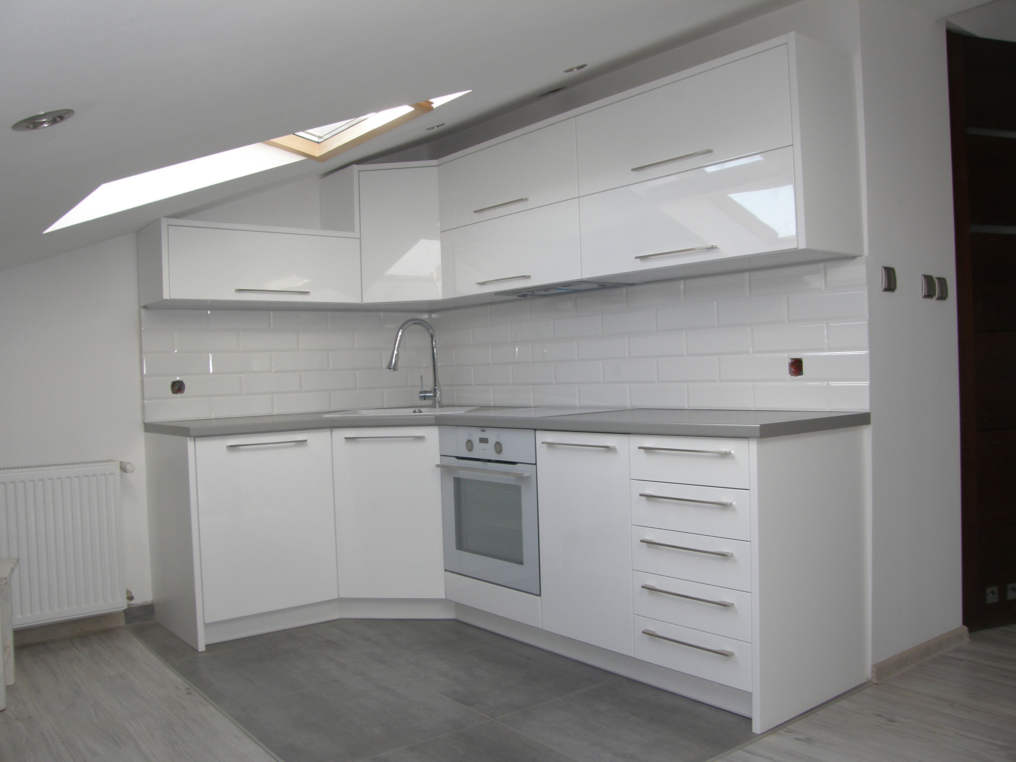 Jasna kuchnia z białymi, lakierowanymi frontami, szarym blatem i białymi płytkami w kształcie cegiełek, oświetlona światłem z okna dachowego.