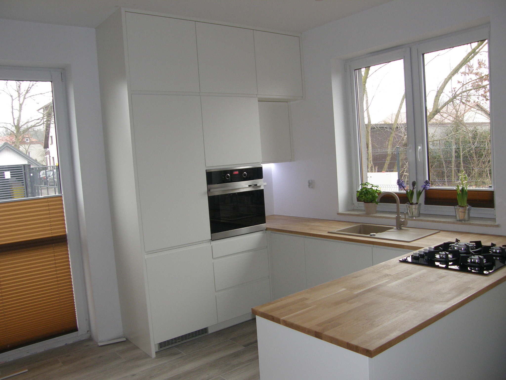 Nowoczesna, jasna kuchnia z białymi frontami szafek, drewnianym blatem i wbudowanym piekarnikiem. Na blacie kuchennym ustawiona płyta gazowa i doniczki z kwiatami przy oknie.
