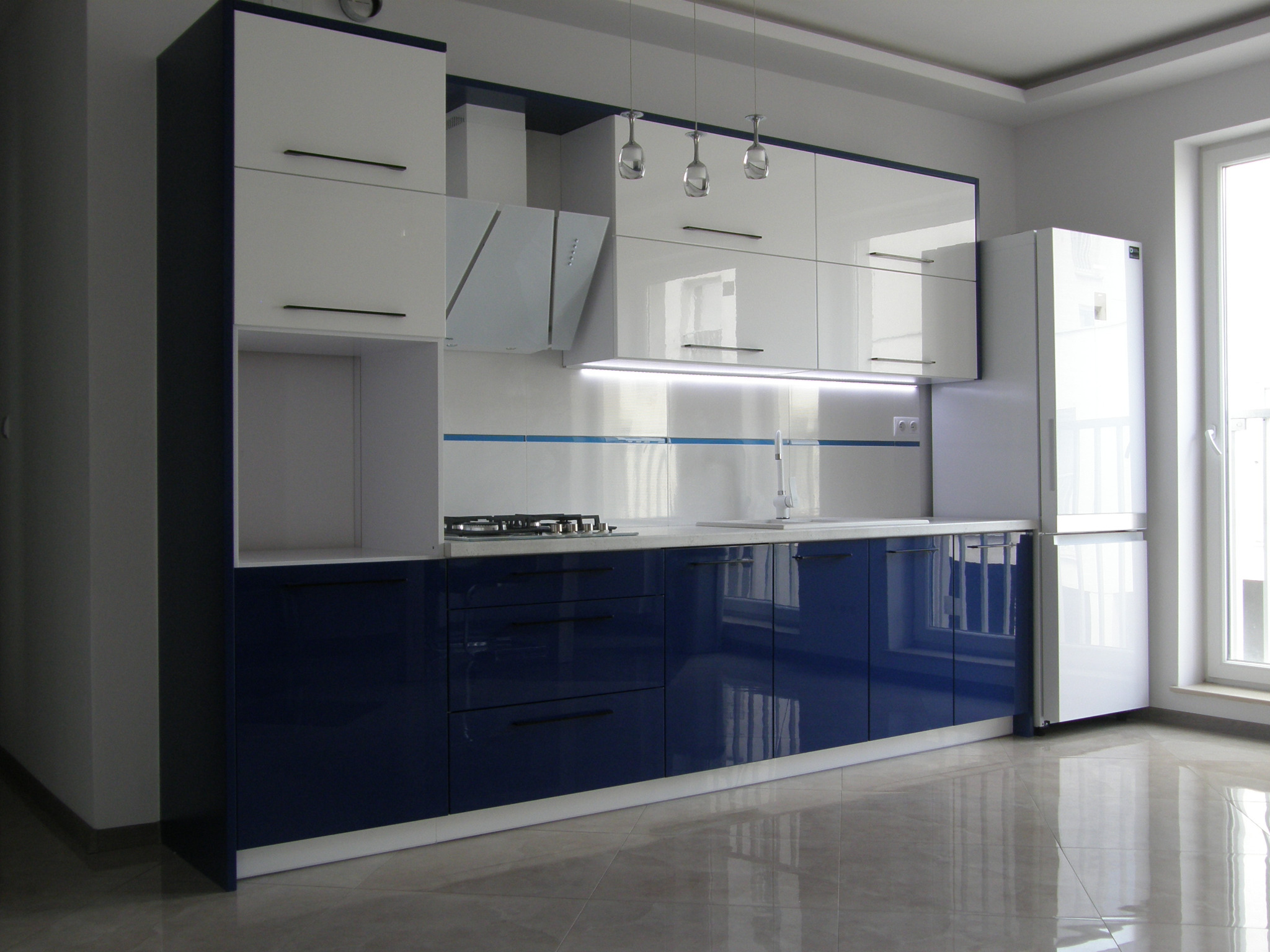 Nowoczesna kuchnia z białymi szafkami górnymi i granatowymi dolnymi, oświetlenie podszafkowe LED, trzy szklane lampy wiszące nad blatem, widok na błyszczącą podłogę.