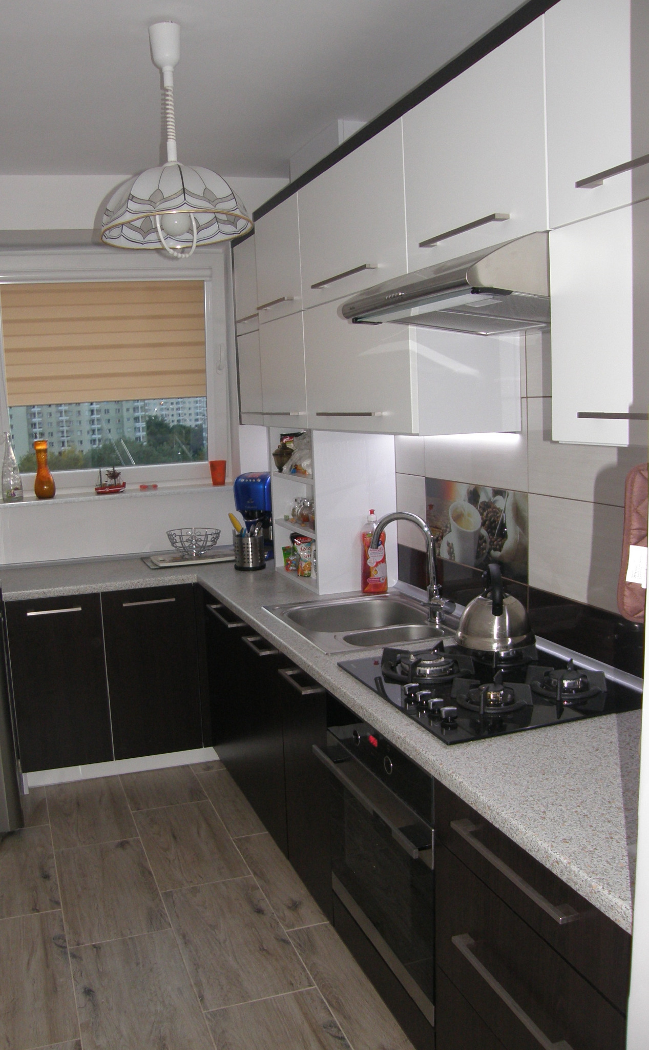 Nowoczesna kuchnia z białymi szafkami górnymi i ciemnobrązowymi dolnymi, szarym blatem, płytą gazową i piekarnikiem, widok na okno z roletą i zabudowę miejską