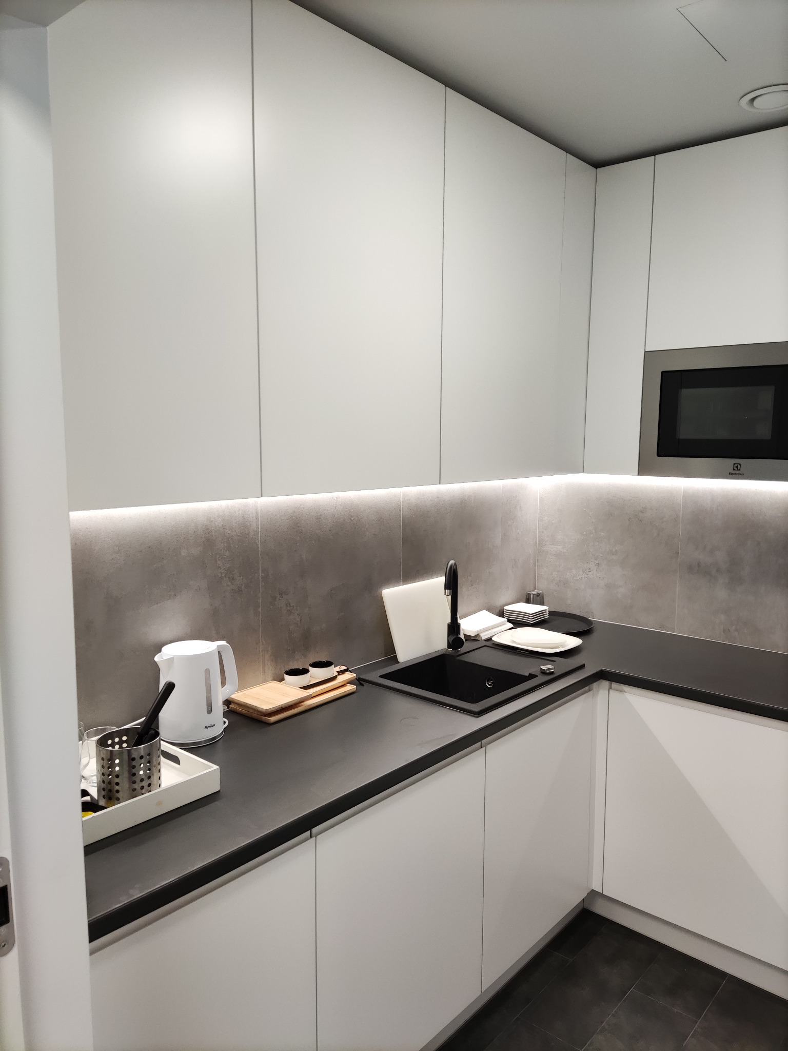 Minimalistyczna, biała zabudowa kuchenna z czarnym blatem i zlewozmywakiem, oświetlenie LED pod szafkami, widoczny czajnik elektryczny Amica i wbudowany piekarnik Electrolux.