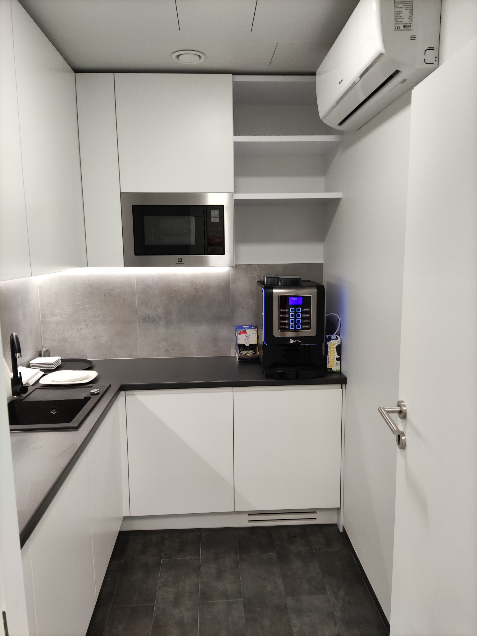 Nowoczesna, minimalistyczna kuchnia z białymi szafkami, czarnym blatem i płytkami imitującymi beton, z wbudowaną mikrofalówką Electrolux i automatycznym ekspresem do kawy Necta. Widoczny...