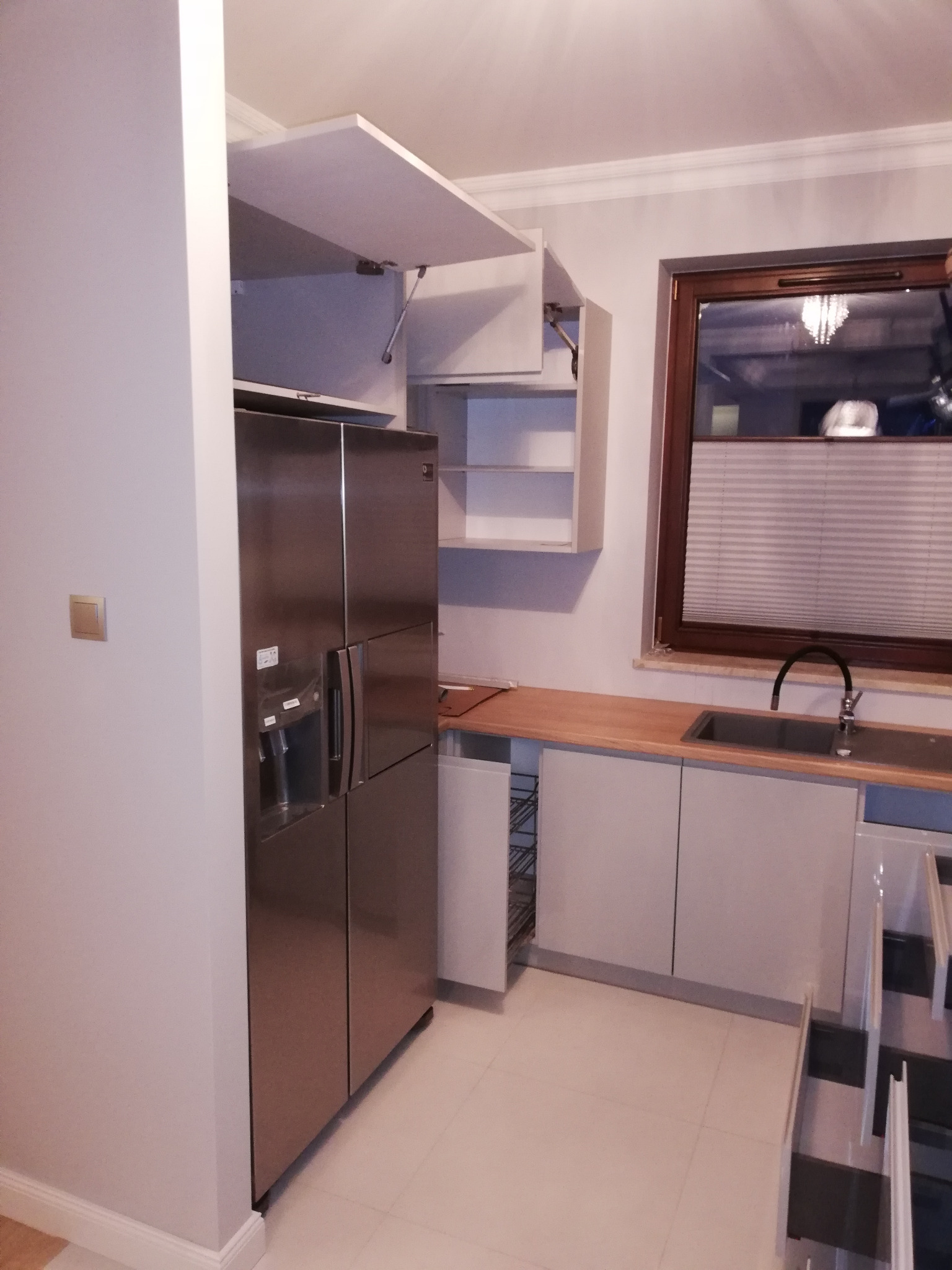 Nowoczesna kuchnia z szarymi szafkami, drewnianym blatem, dużym, srebrnym lodówko-zamrażarką i otwartą szafką górną z podnośnikiem gazowym.