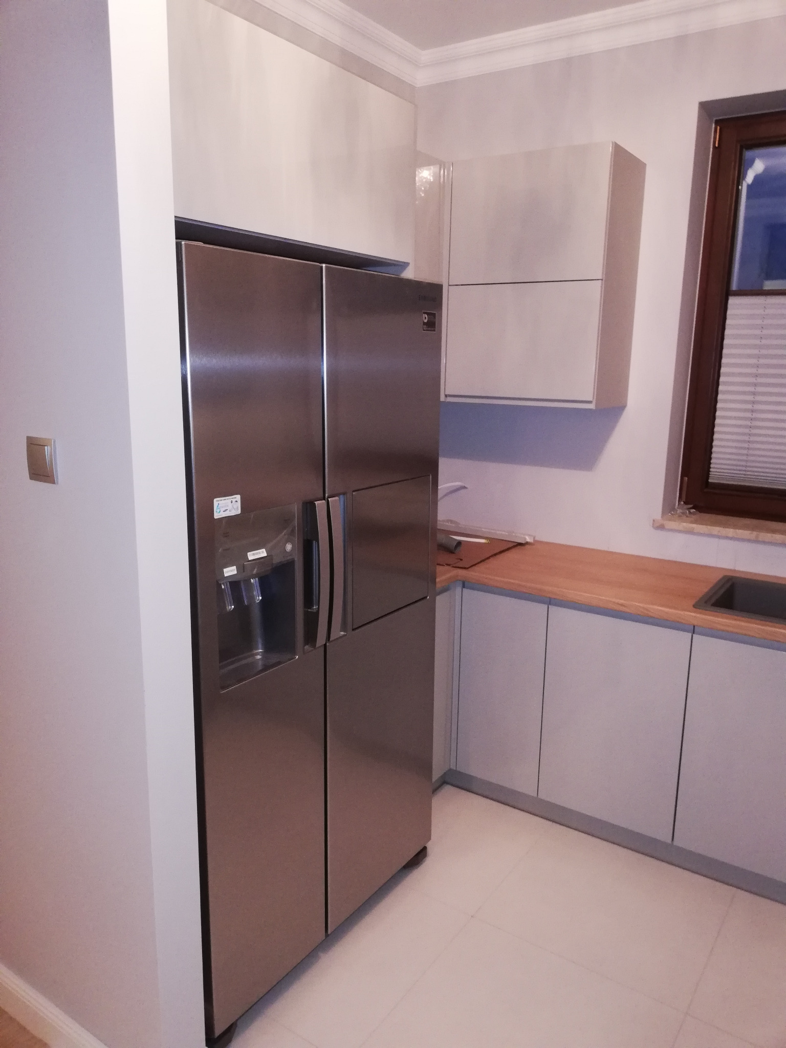 Nowoczesna kuchnia z szarymi szafkami, drewnianym blatem i dużą, srebrną lodówką z kostkarką do lodu.