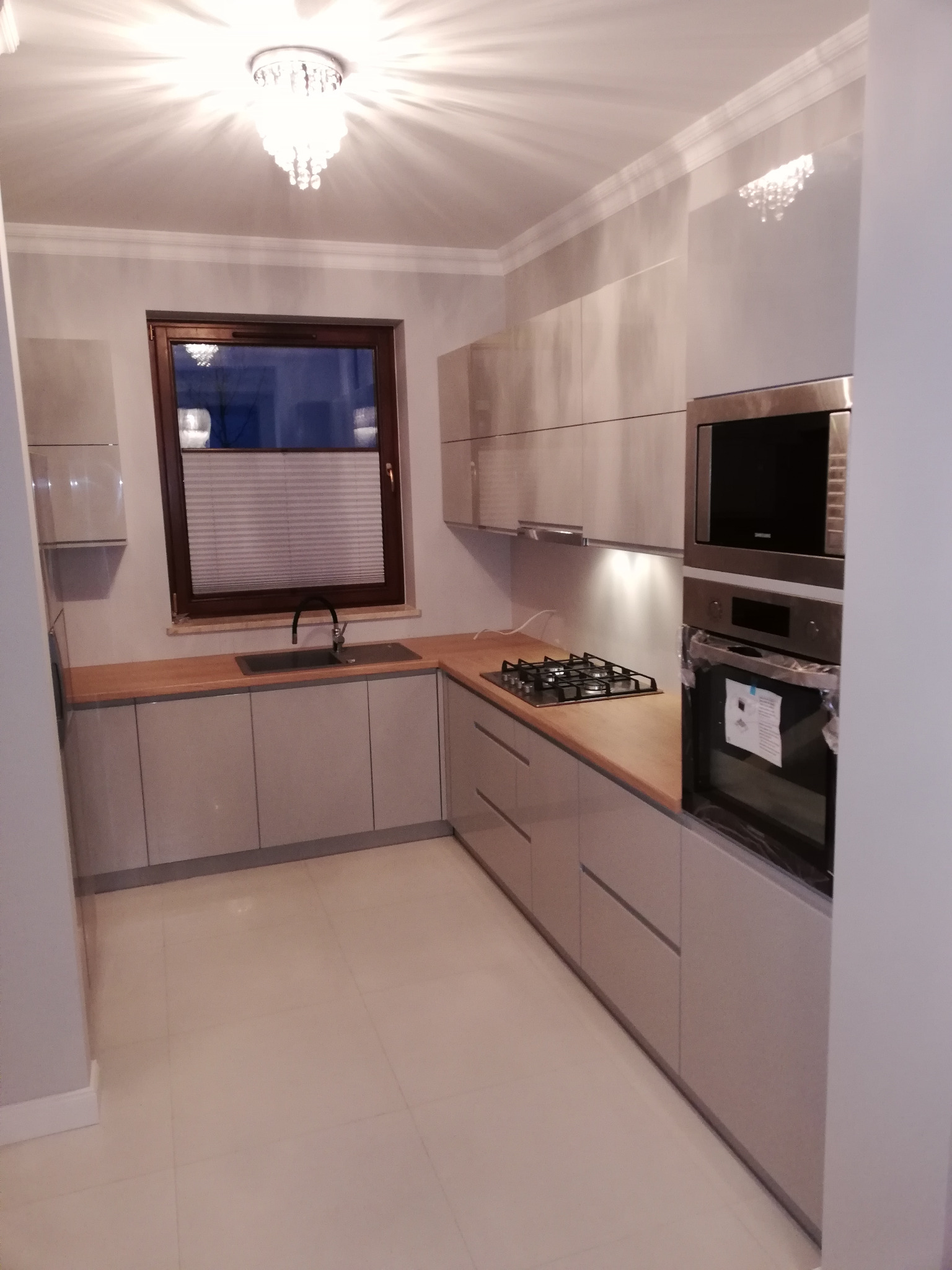 Nowoczesna kuchnia z szarymi frontami, drewnianym blatem, płytą gazową i piekarnikiem w zabudowie, oświetlona kryształowym żyrandolem.