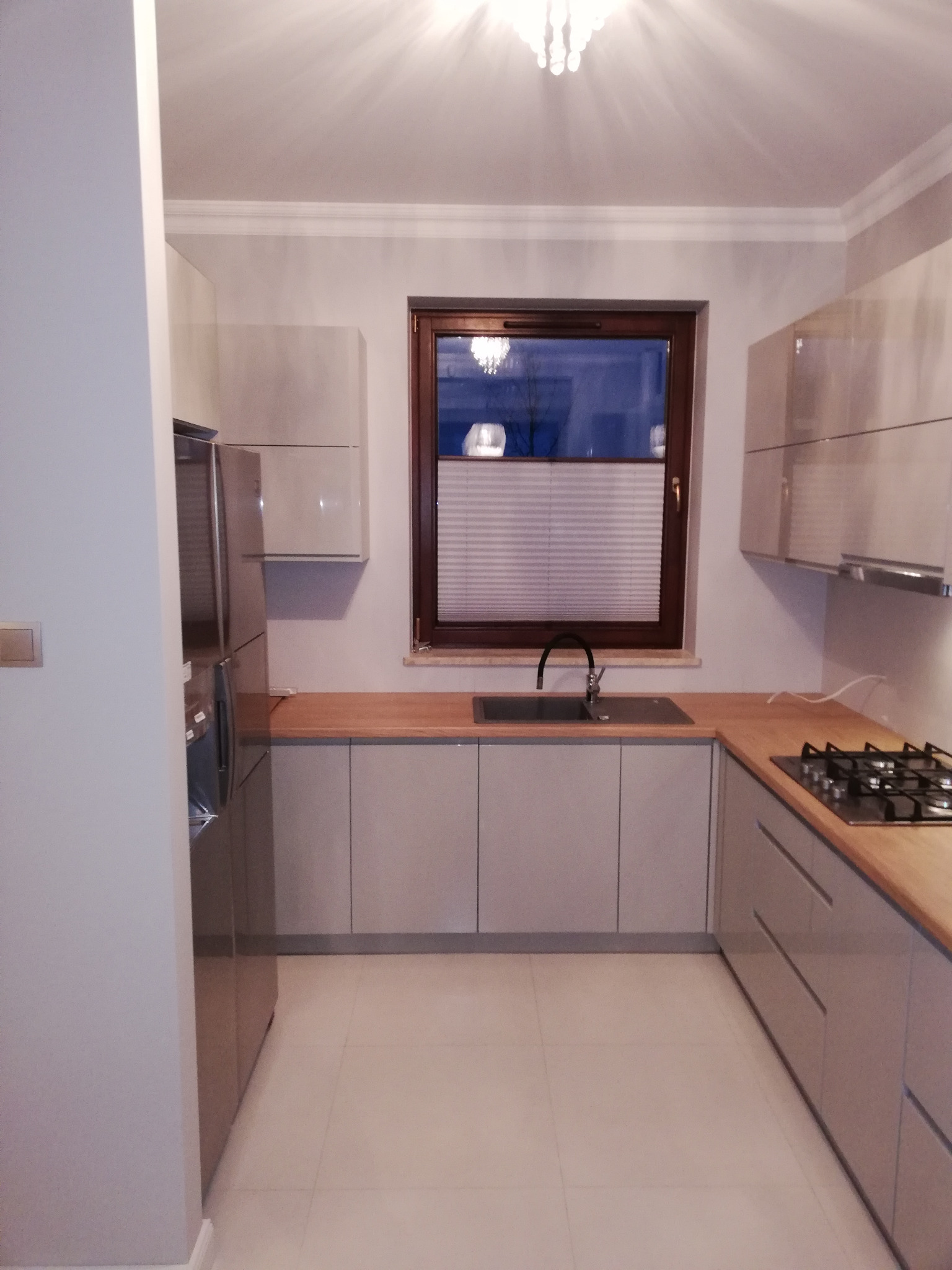 Jasna kuchnia z drewnianym blatem, widok na okno z roletami plisowanymi, srebrna lodówka po lewej stronie.