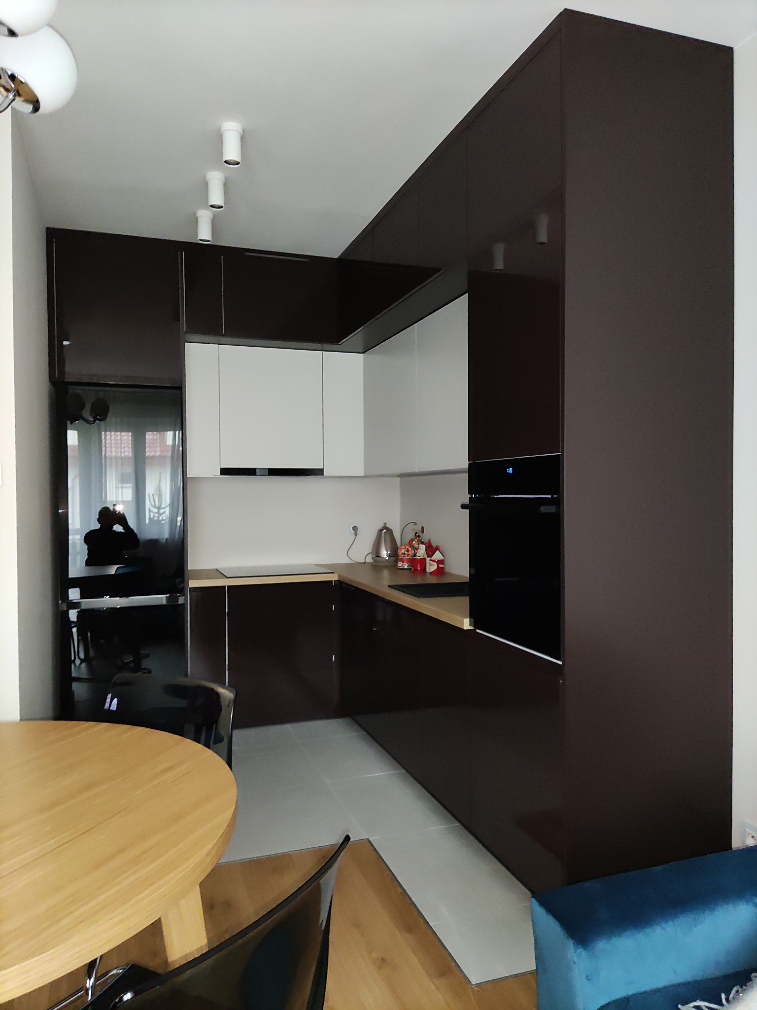 Nowoczesna kuchnia z brązowymi, lakierowanymi szafkami, białymi górnymi szafkami i drewnianym blatem, oświetlona punktowymi lampami sufitowymi, z odbiciem fotografa w czarnym froncie lodówki.