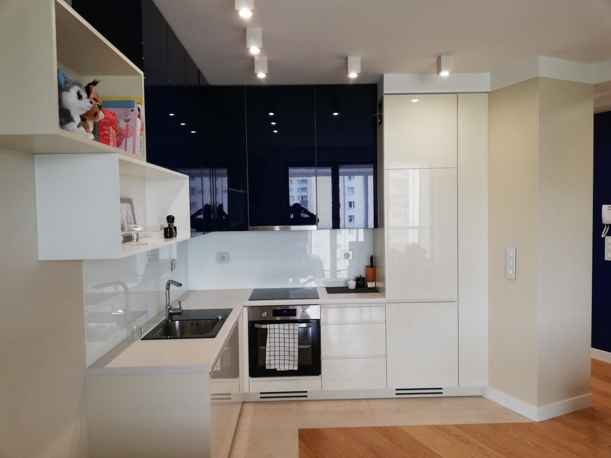 Nowoczesna kuchnia z białymi i granatowymi szafkami na wysoki połysk, szklanym panelem nad blatem, piekarnikiem z ręcznikiem i półką z dekoracjami.