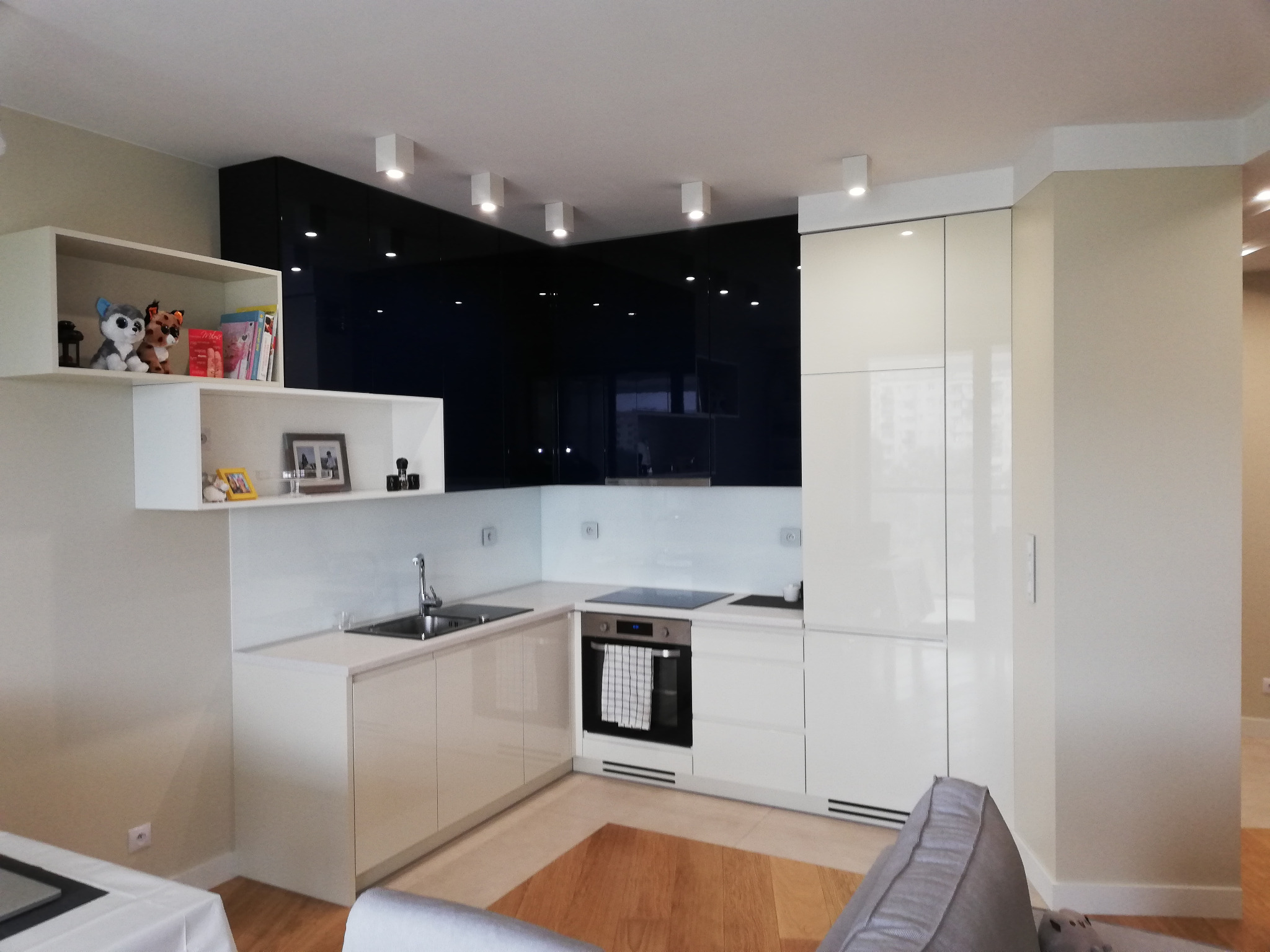 Nowoczesna kuchnia z białymi i czarnymi frontami, oświetleniem punktowym i drewnianą podłogą. Na półce dekoracyjne przedmioty, w tle widoczny salon.