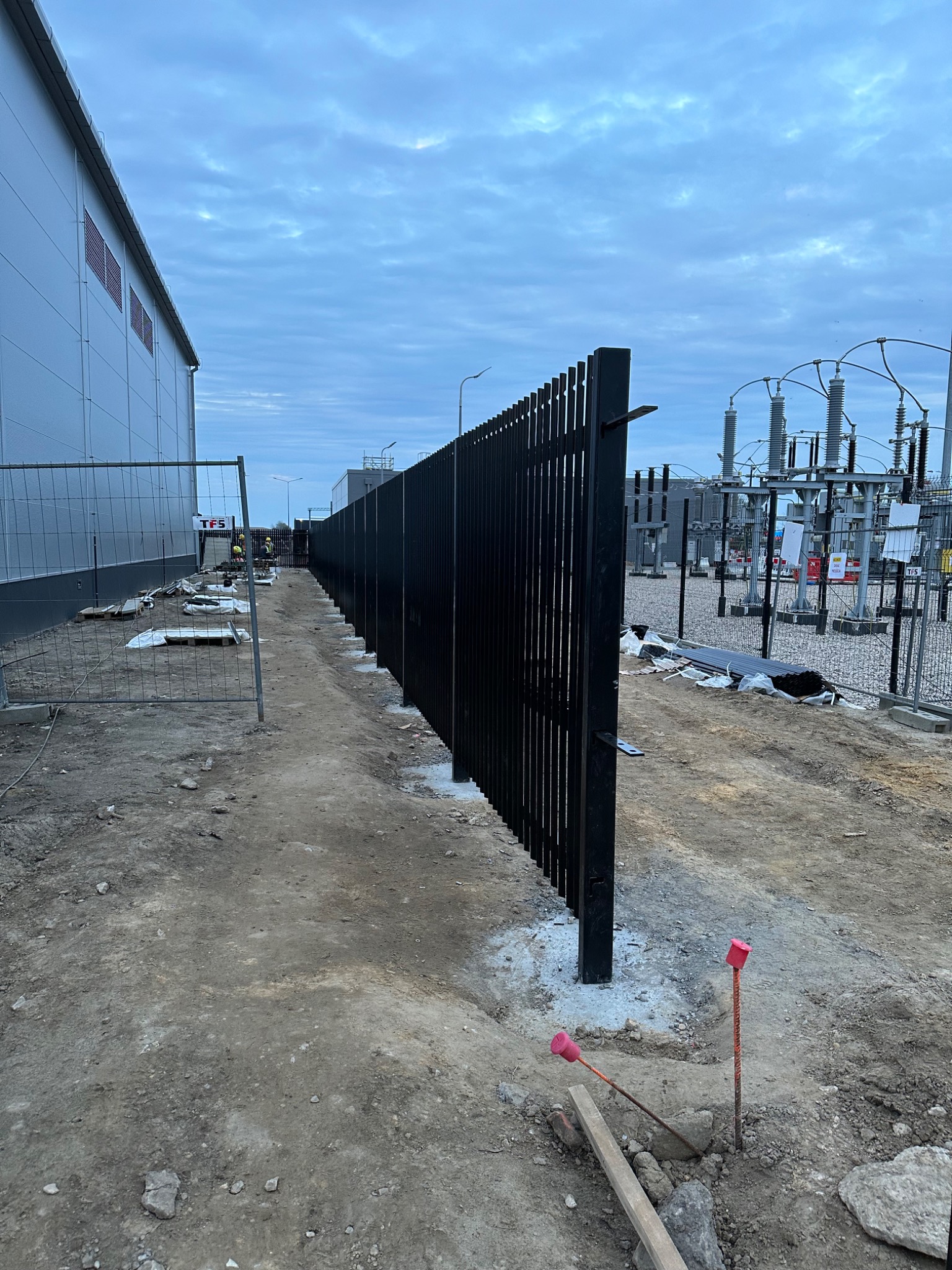Montaż nowoczesnego, czarnego ogrodzenia panelowego na betonowej podmurówce, obok budynku przemysłowego i stacji transformatorowej, widoczne pręty z czerwonymi zaślepkami w gruncie.