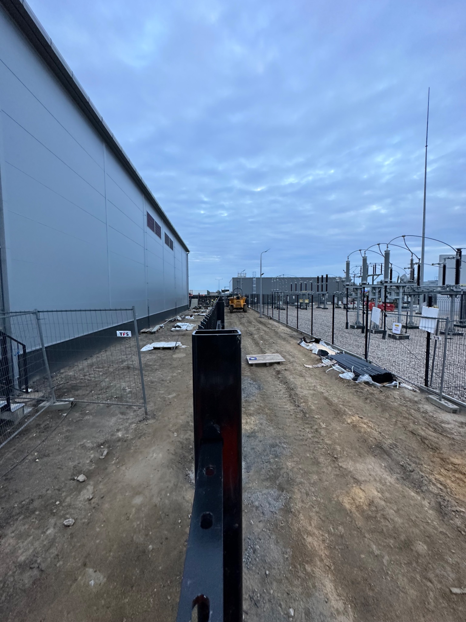 Przygotowanie terenu pod budowę: wyrównana, ubita droga gruntowa z widocznym sprzętem budowlanym w oddali, obok ogrodzenie i budynek przemysłowy.