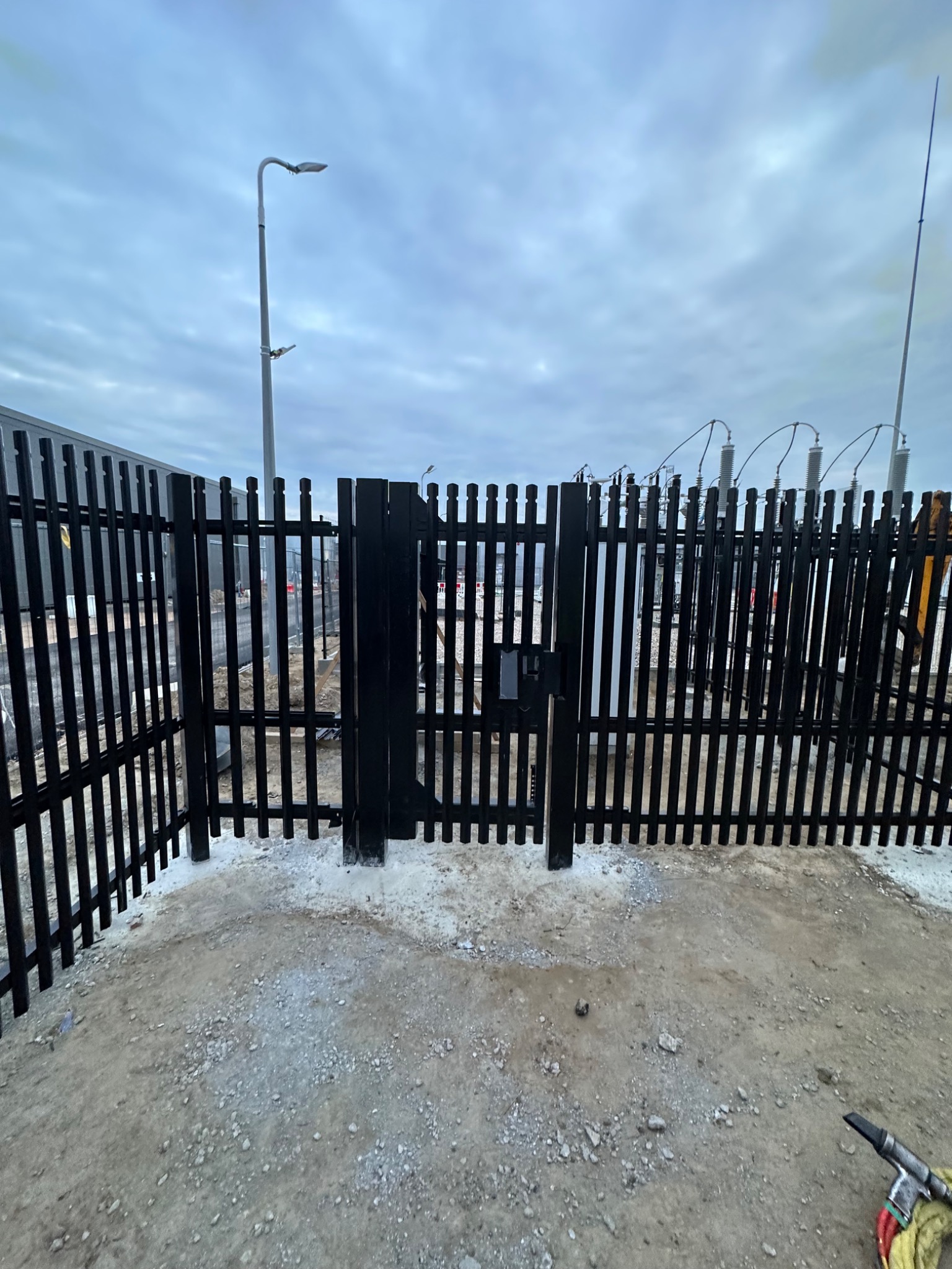 Czarna metalowa brama z furtką na terenie przemysłowym, widoczne słupy energetyczne i lampa uliczna w tle, teren utwardzony szarym żwirem.