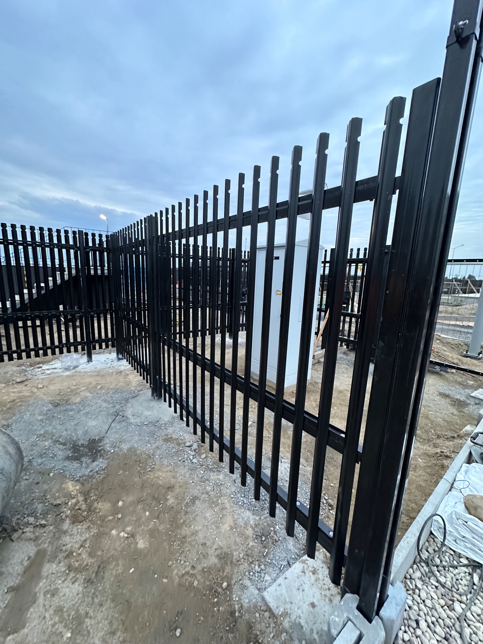 Nowo postawione, czarne, metalowe ogrodzenie panelowe z pionowymi sztachetami, osadzone na betonowych fundamentach, na placu budowy z widoczną skrzynką elektryczną w tle.